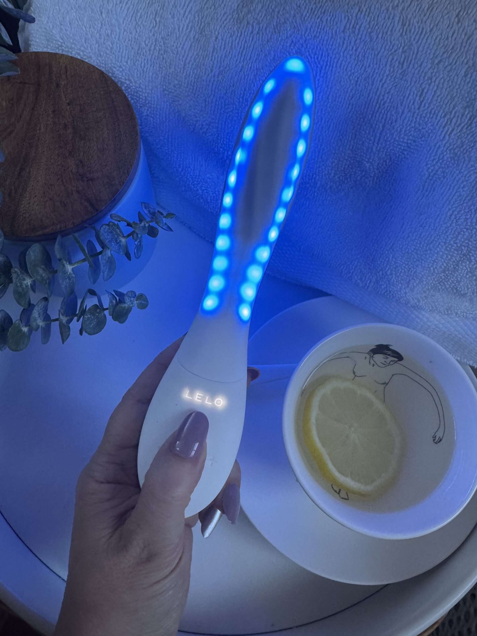 Hand holding a LELO brand LED light therapy wand next to a teacup with a lemon slice.