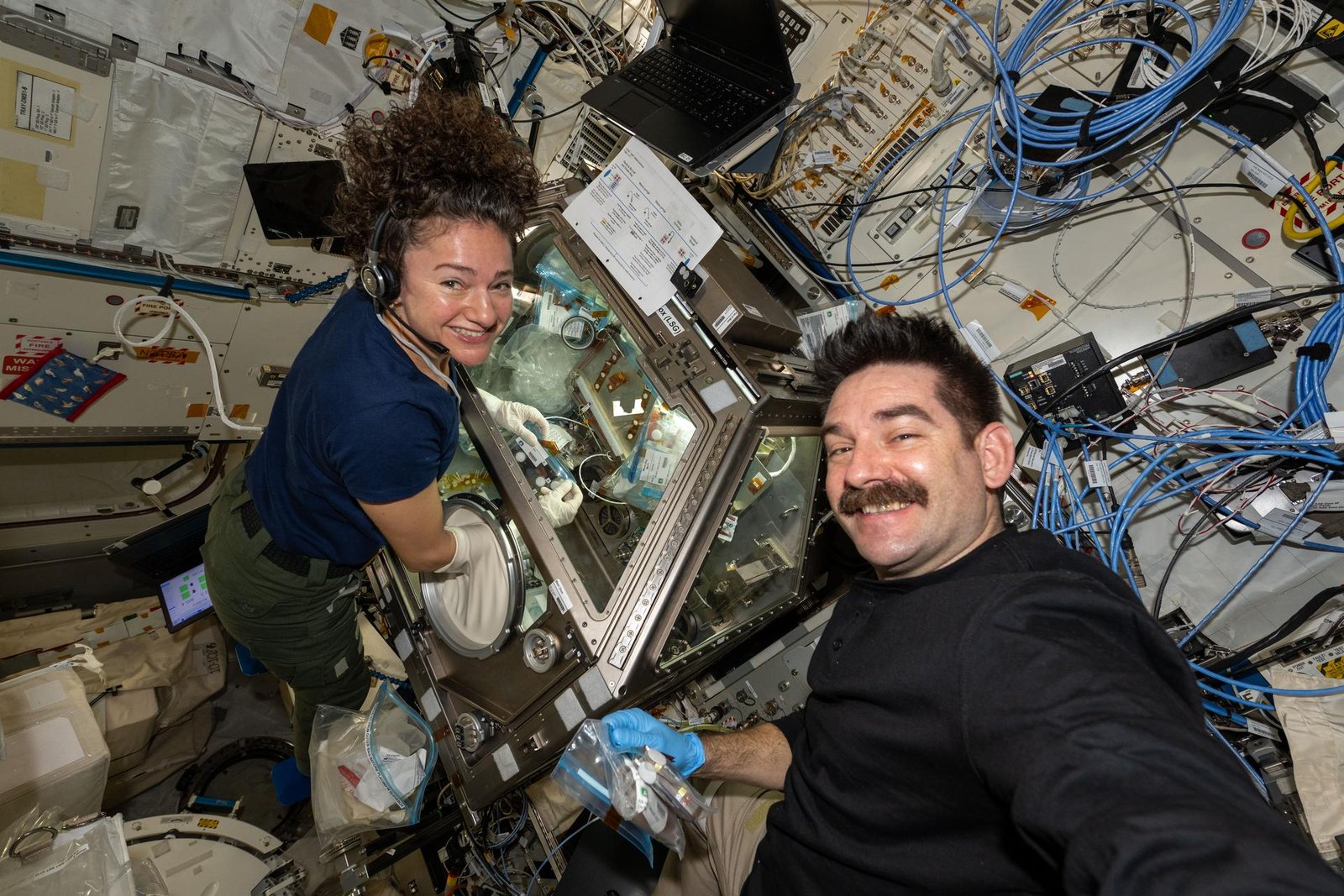 NASA astronauts Jessica Meir (left) and Jack Hathaway (right) smile at the camera as Hathaway takes a selfie aboard the International Space Station. Meir wears blue gloves while performing operations inside the Life Sciences Glovebox, surrounded by metallic panels, cables, and equipment.