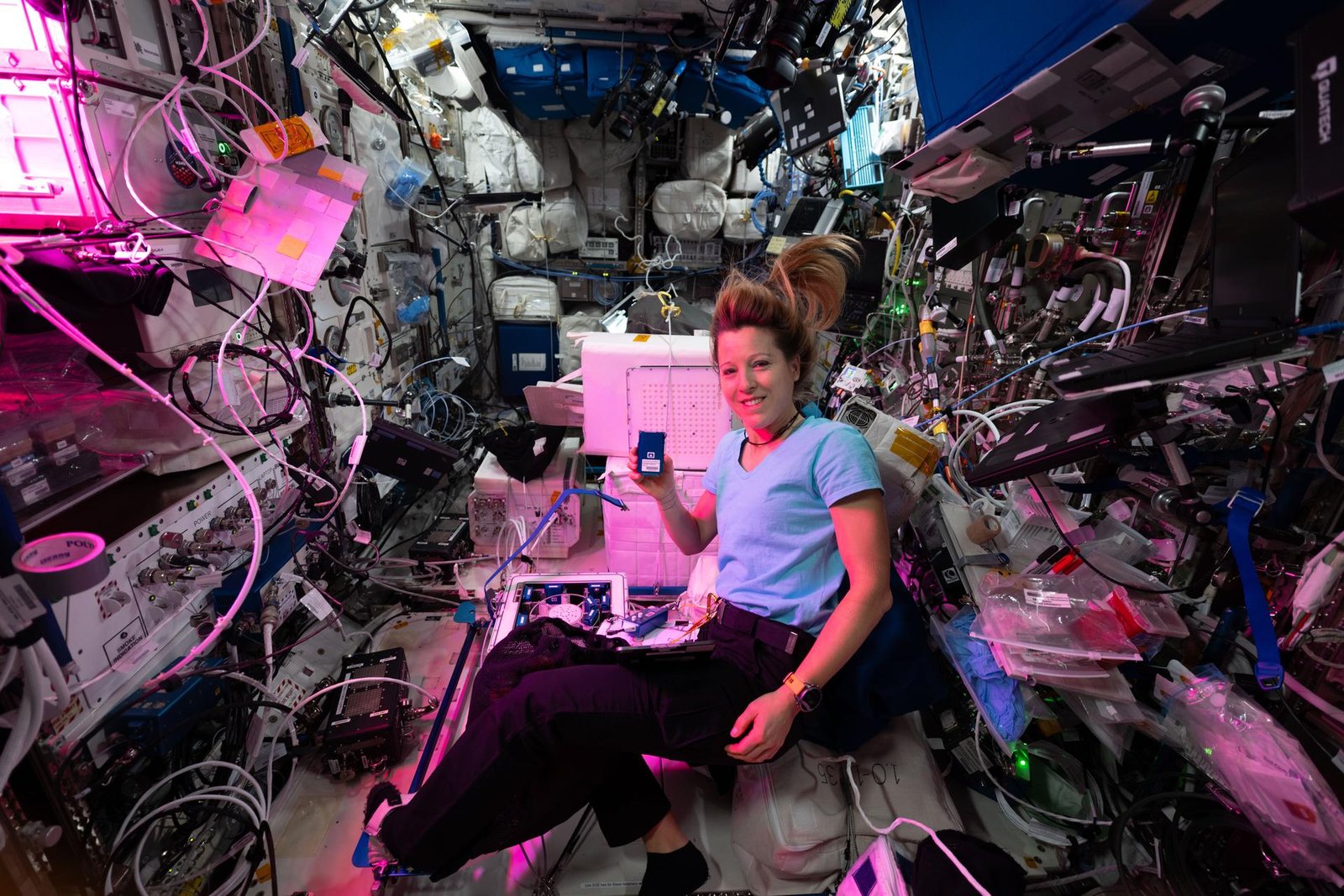 ESA (European Space Agency) astronaut Sophie Adenot floats on the International Space Station, surrounded by wires, compartments, and plastic packages. There is a pink light emanating from the left side of the image and it shines on Sophie and the objects around her. She smiles and holds up a blue, metallic rectangle in her hand.