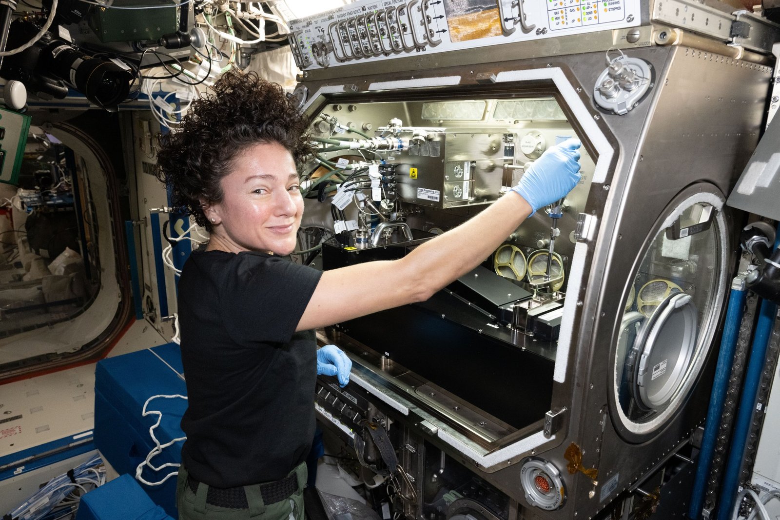 NASA astronaut Jessica Meir smiles at the camera in front of the rectangular Microgravity Science Glovebox aboard the International Space Station. Wearing blue gloves, she reaches into the open compartment, which contains experiment hardware surrounded by metallic panels, cables, and tubing.