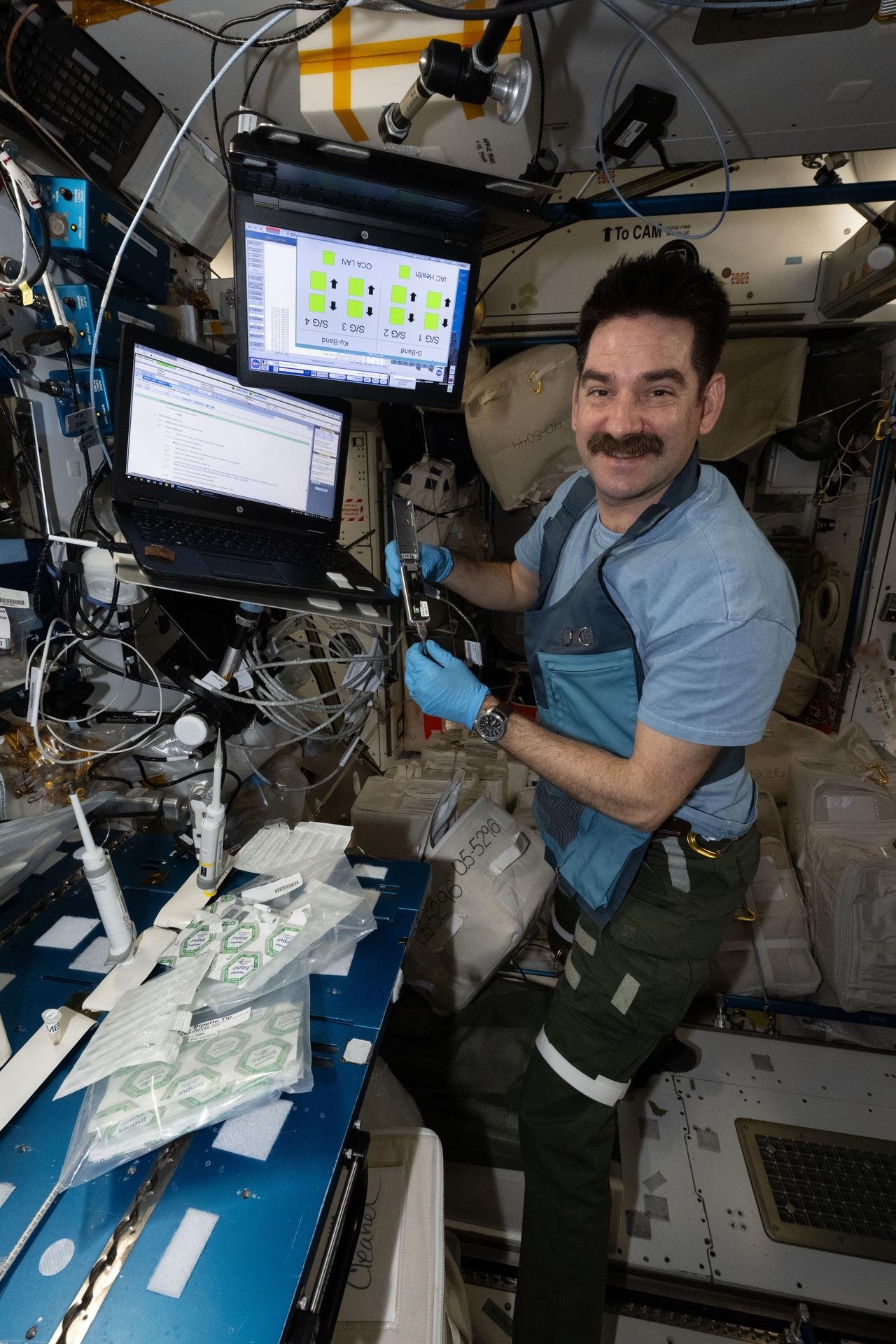 NASA astronaut Jack Hathaway works aboard the International Space Station. He is wearing blue gloves while holding a metallic device with a hinge at its center. To his left are several plastic packages and a pipette, with two laptop screens mounted above the work area.