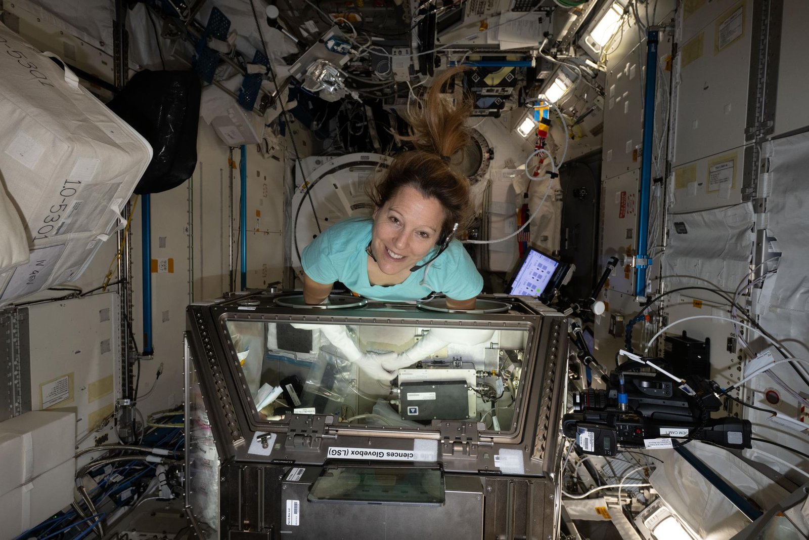 ESA (European Space Agency) astronaut Sophie Adenot smiles at the camera as she reaches inside the large, rectangular Life Sciences Glovebox aboard the International Space Station. Her ponytail floats above her head as she leans into the open compartment, surrounded by reflective silver panels, cables, tubing, and equipment.