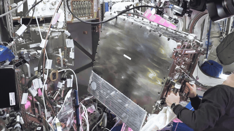 ESA (European Space Agency) astronaut Sophie Adenot carefully guides the Metal 3D Printer into place aboard the International Space Station. She uses both hands to position the large, enclosed, rectangular printer into its slot, surrounded by cables and other station hardware.