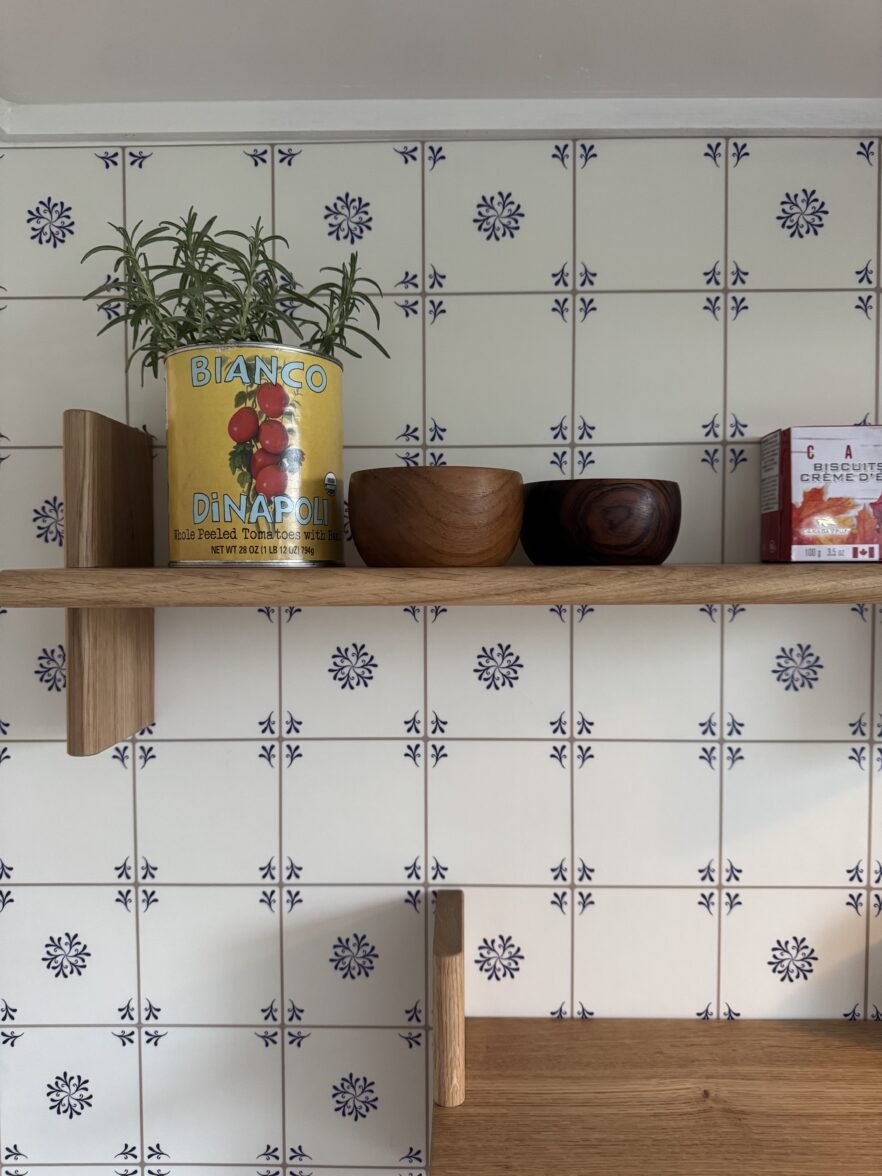 A wooden shelf against a blue and white patterned tile wall holds a Bianco DiNapoli tomato can containing a small plant, two wooden bowls, and a box of biscuits.