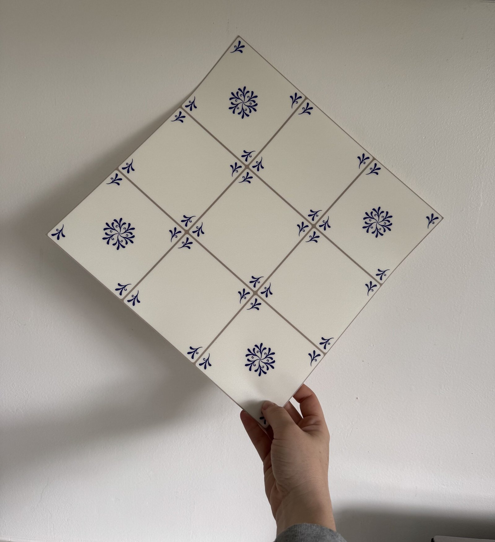 A hand holds a large ceramic tile composed of nine smaller white tiles with blue floral patterns, arranged in a 3x3 grid.