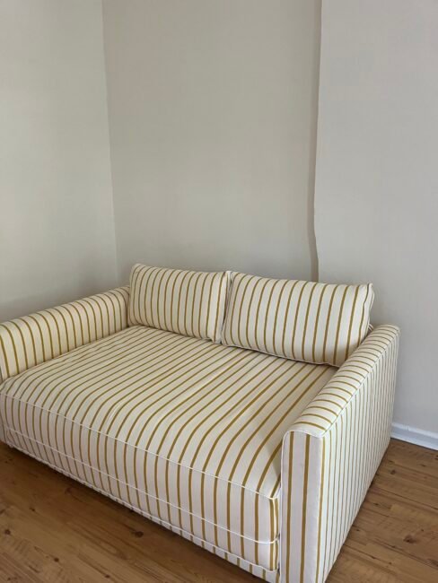 A striped yellow and white sofa with two back cushions sits on a wooden floor against a plain beige wall.