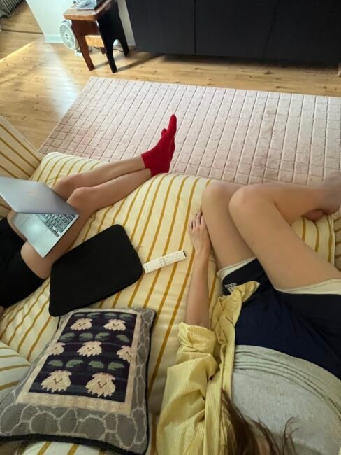 Two people relax on a striped couch; one uses a laptop, and a remote control rests on the cushion between them. A pillow and a laptop sleeve are also visible.