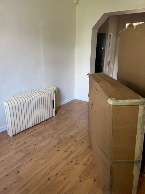 A room with a wooden floor, a striped cushion or mattress leaning against the wall, and a large cardboard box standing upright near the center.