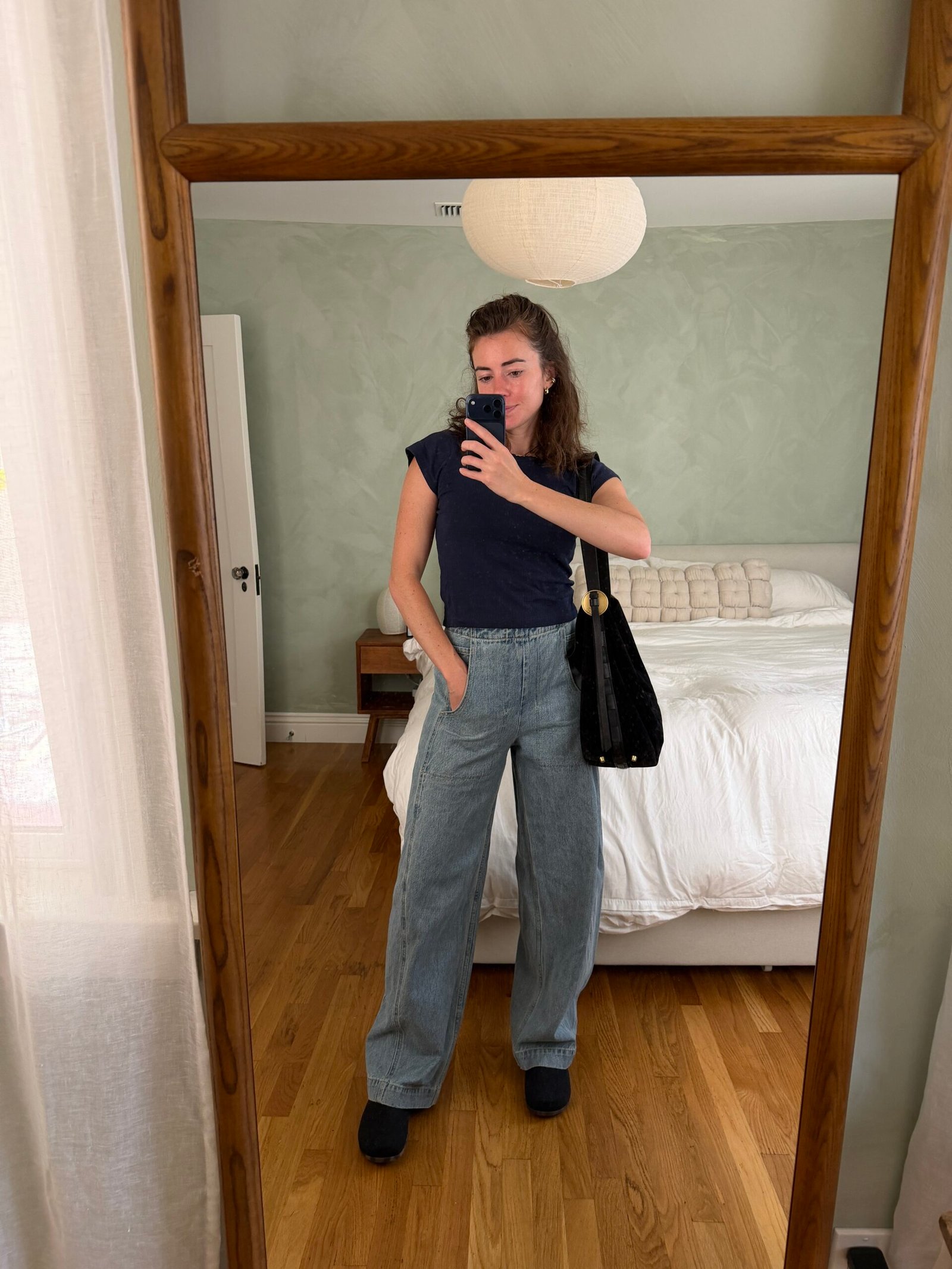 A person takes a mirror selfie wearing a dark blue t-shirt, wide-leg jeans, and a shoulder bag.
