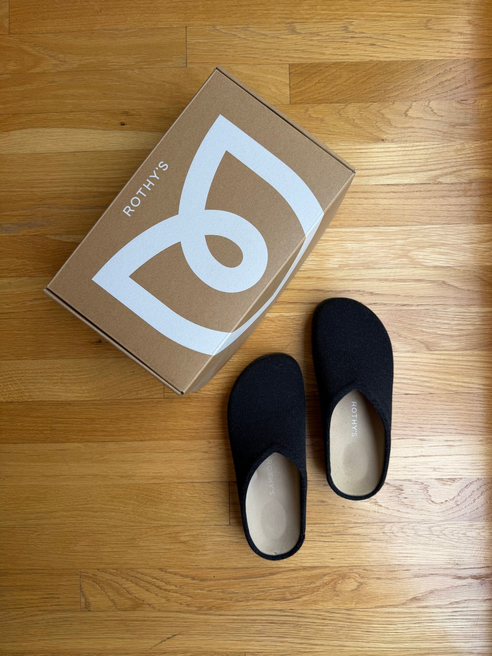 A pair of black Rothy’s slip-on shoes next to their branded cardboard box on a wood floor.
