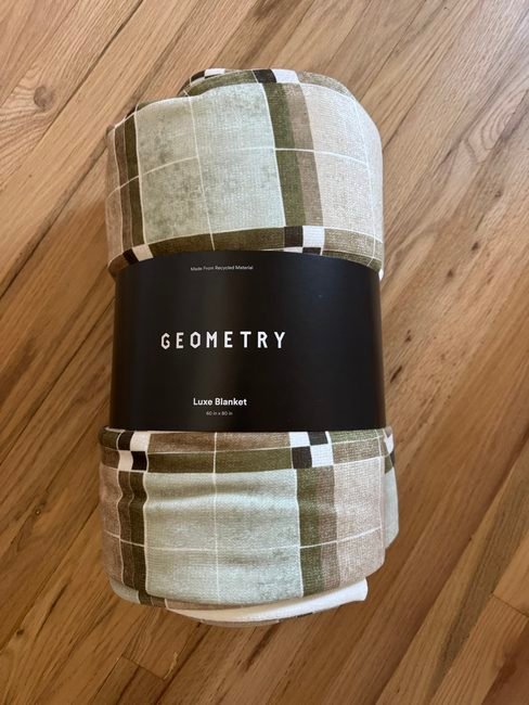 A rolled, plaid-patterned Geometry Luxe Blanket with a product band reading “Made From Recycled Material,” “GEOMETRY,” and “60 in x 80 in.”
