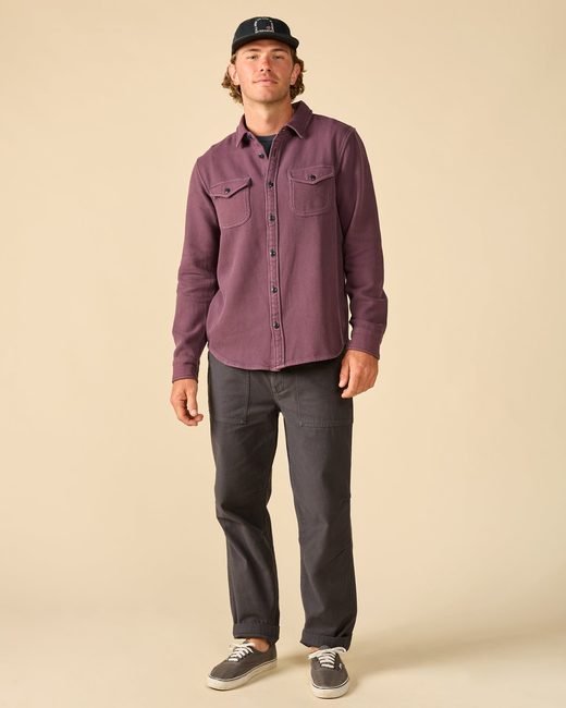 A man wearing a black baseball cap, a plum button-down shirt, dark gray pants, and gray sneakers.