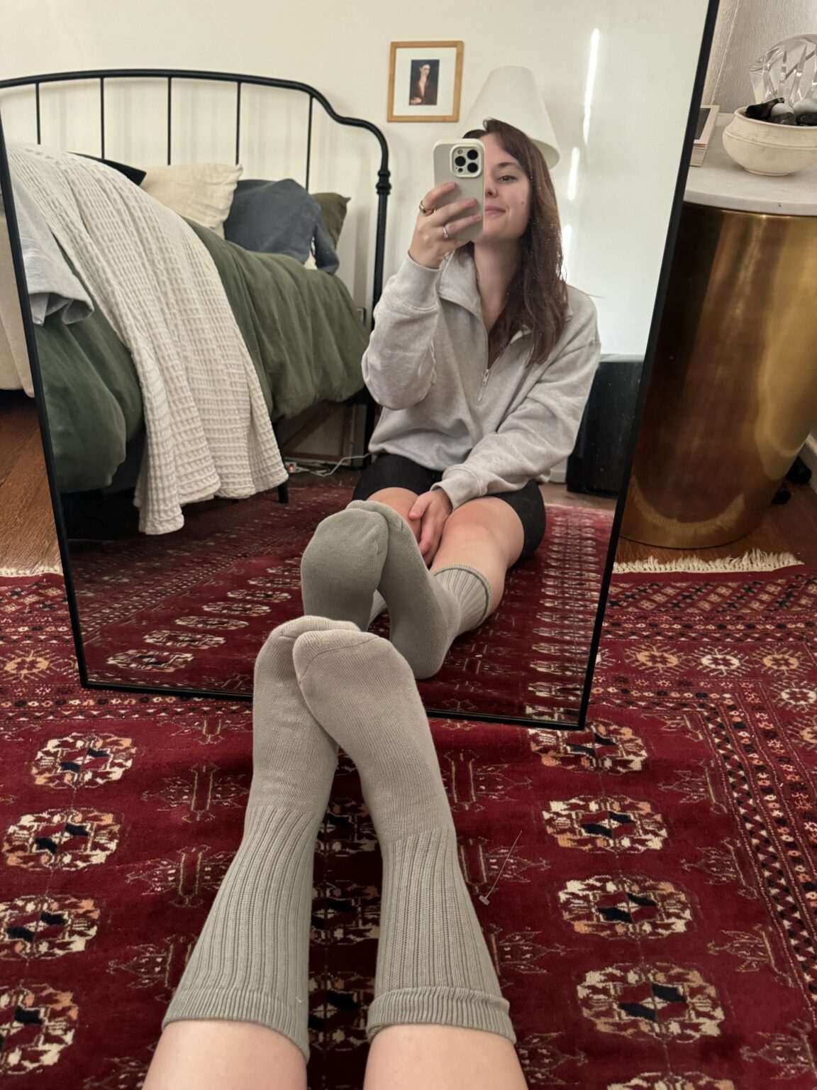 A person sits on a patterned rug, taking a mirror selfie while wearing gray ribbed socks.