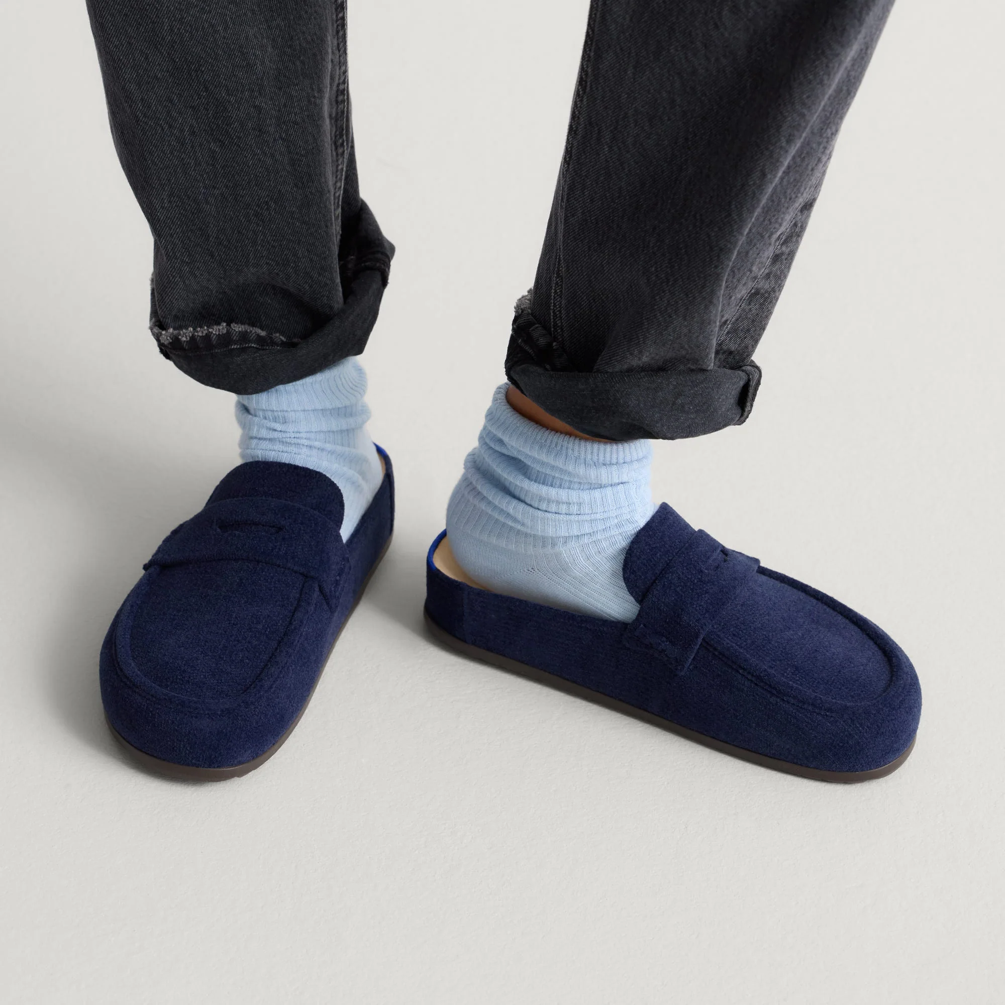 Person wearing dark jeans, light blue socks, and navy blue suede mule loafers.