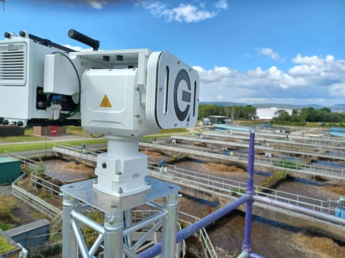 White monitoring instrument mounted above wastewater treatment tanks, with metal walkways, railings and other treatment infrastructure in the background.
