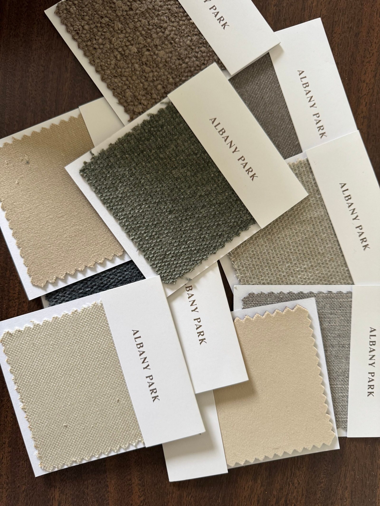 A scattered arrangement of Albany Park fabric swatches in various neutral tones, including beige, gray, and taupe, displayed on a dark wood surface.