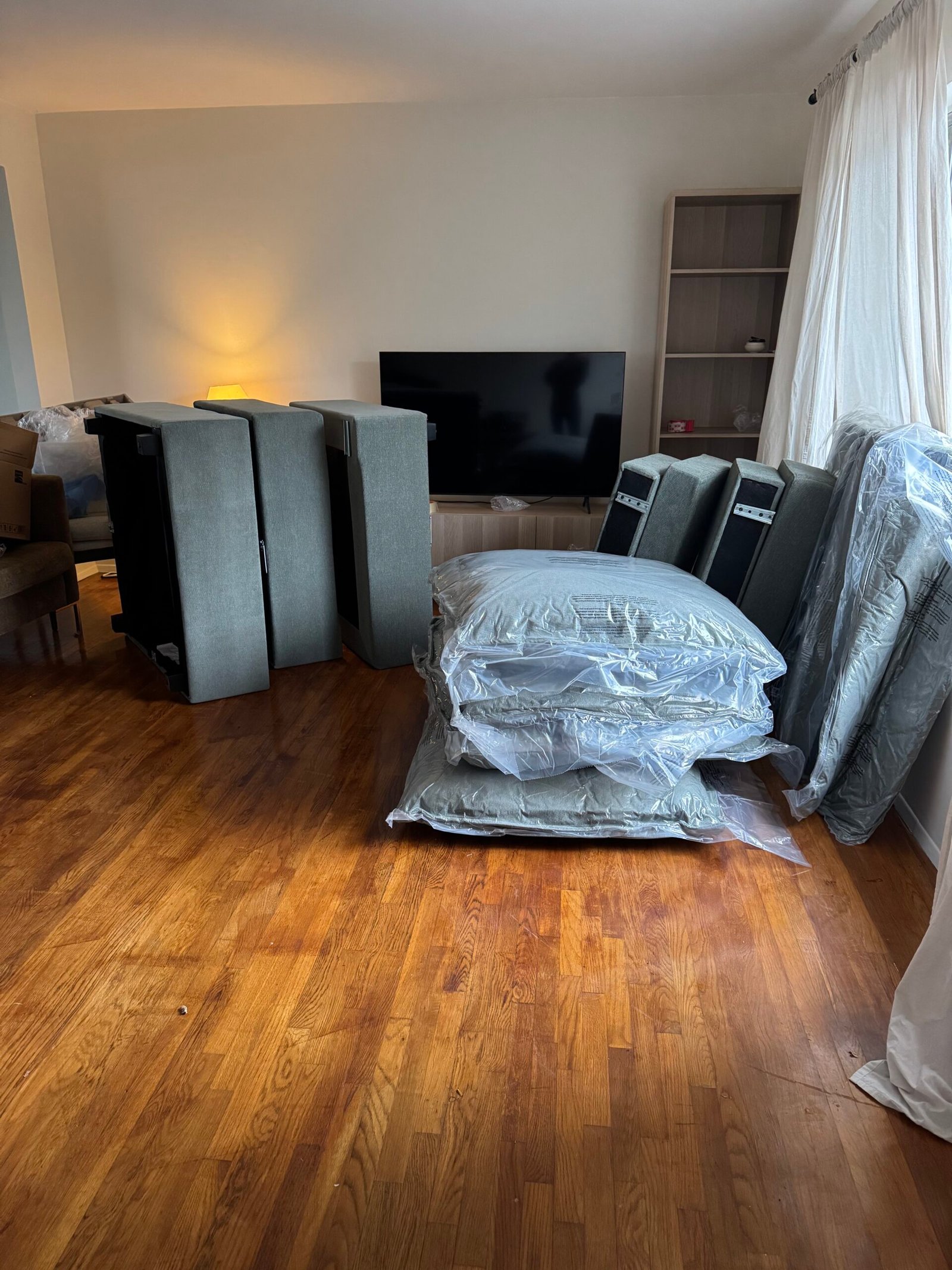 Couch cushions and parts wrapped in plastic are arranged on a wooden floor in a living room, with a TV and empty shelves in the background.