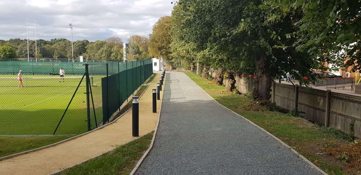 Outdoor tennis courts enclosed by green fencing, with a gravel pathway running alongside mature trees and a narrow grass verge