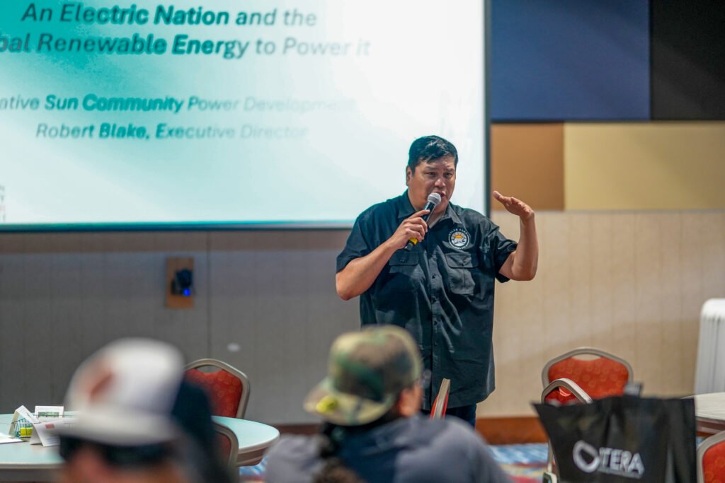 Man speaking into a microphone in a conference room standing in front of a projector screen.