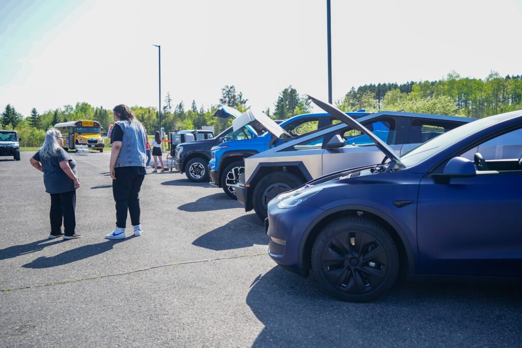 Row of EVs with open frunks in a parking lot with two individuals pictured chatting next to them.