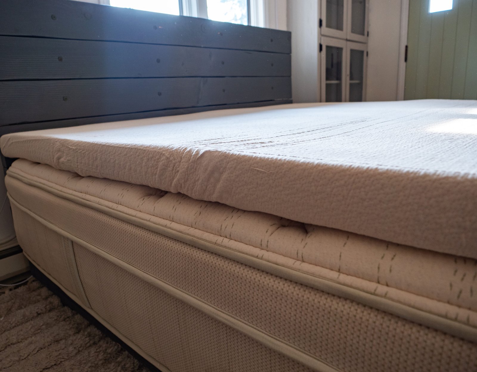 A close-up view of a mattress with a foam mattress topper in a sunlit bedroom, showing the bed frame and part of the room's furniture.
