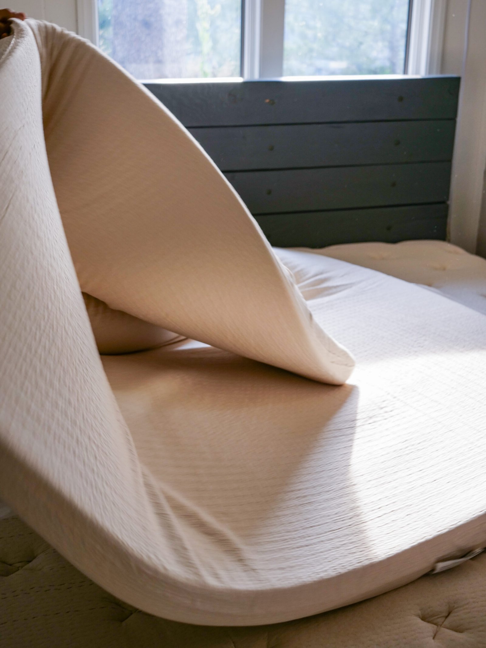 A mattress topper is partially folded back, revealing the mattress underneath, in a sunlit bedroom with a window and dark headboard.