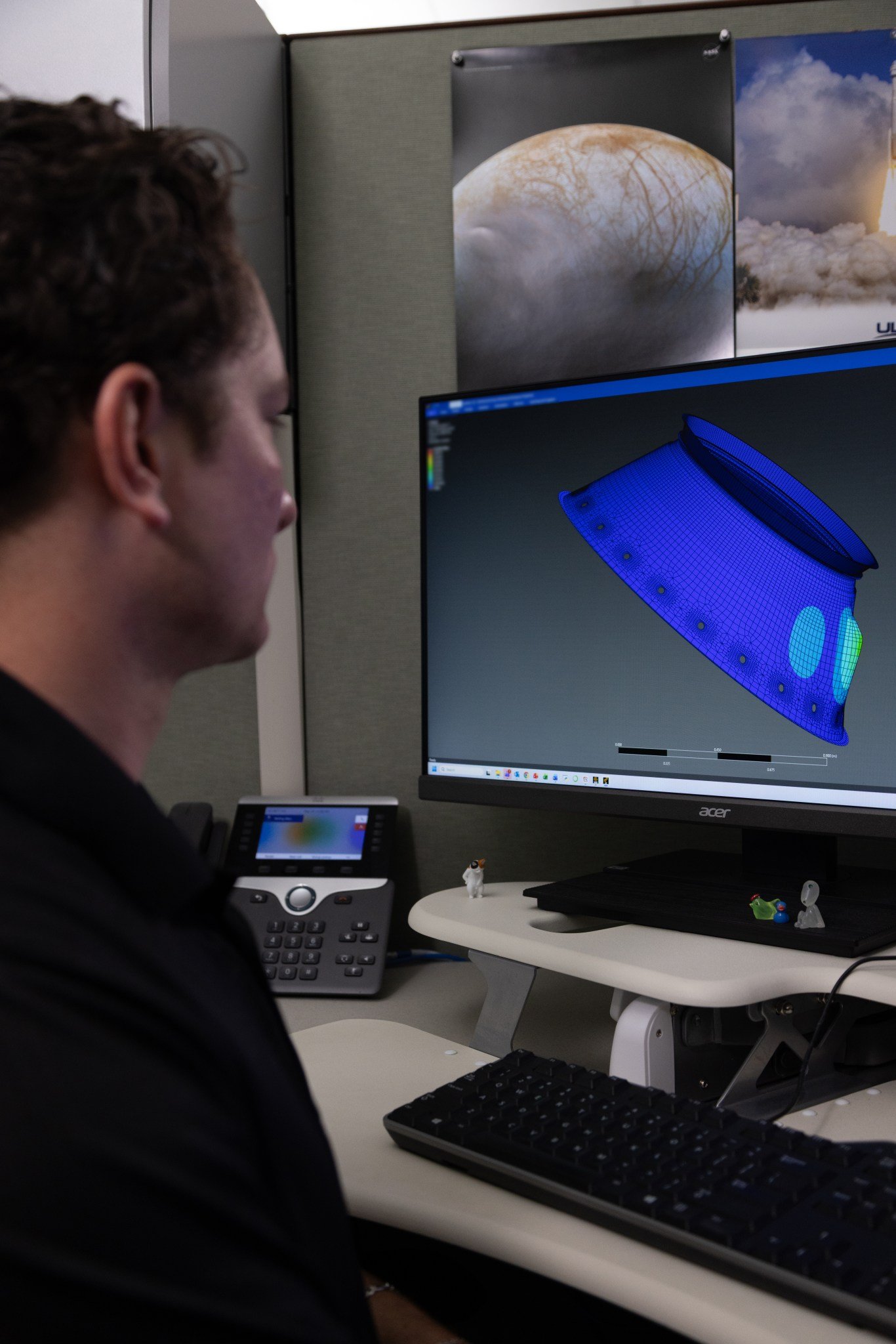 A person sits at a workstation viewing a computer monitor that displays a blue 3D wireframe engineering model of a spacecraft component.