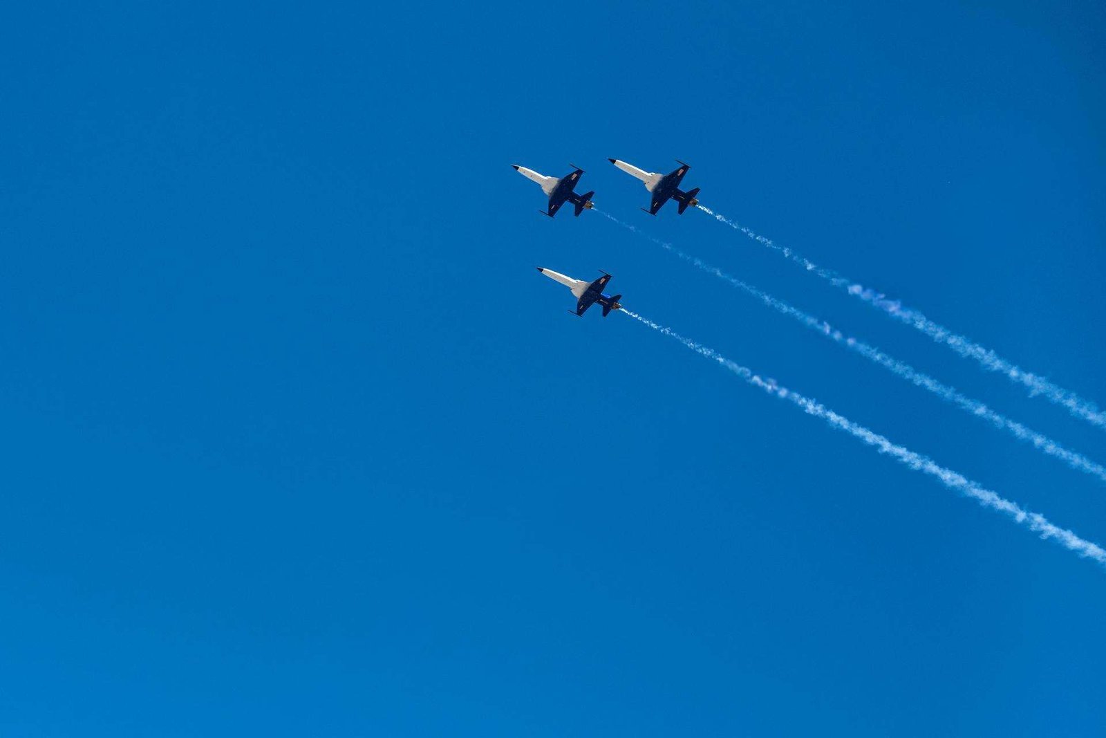 Looking up, three white and black fighter jets fly in formation across a bright, blue sky.