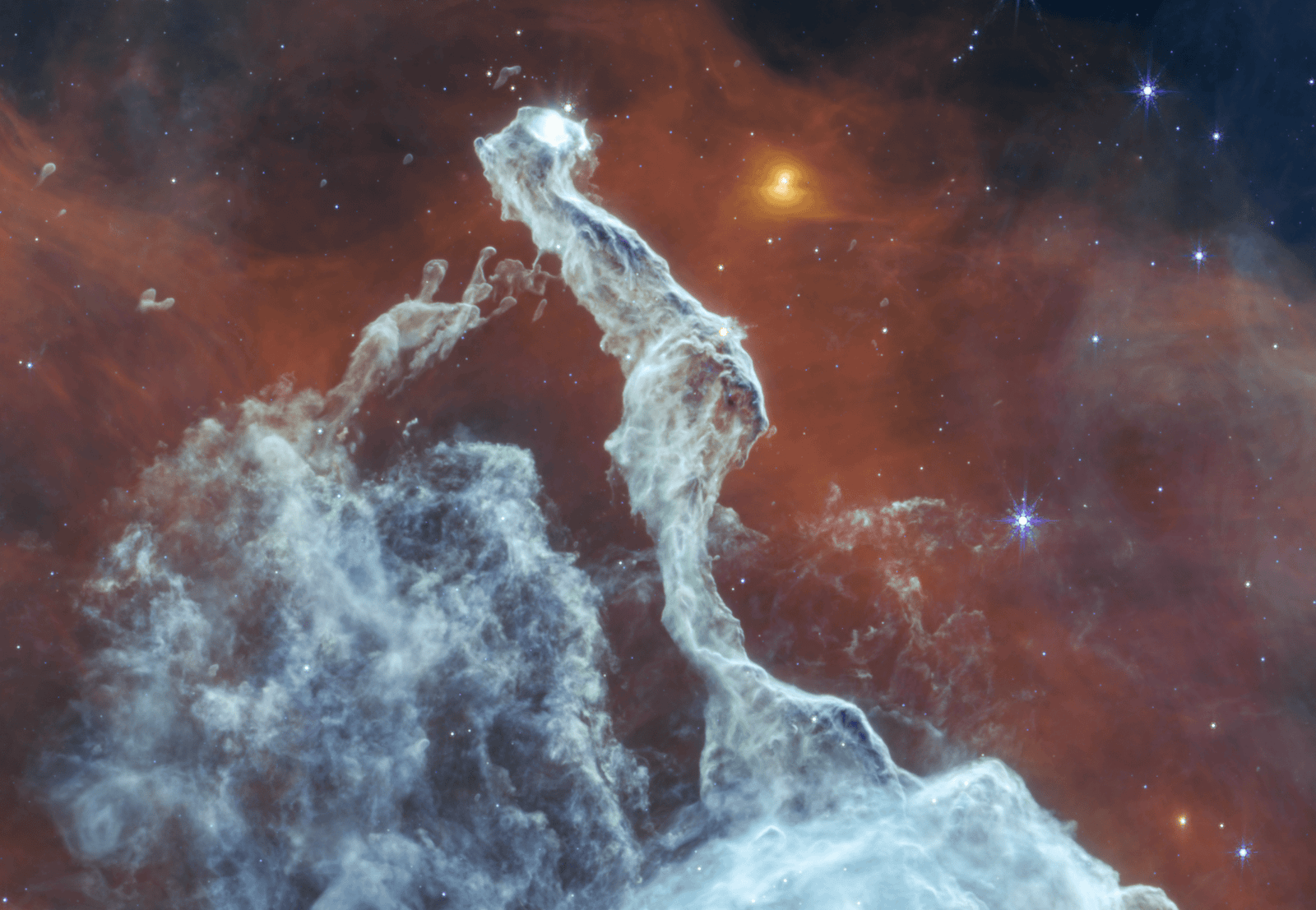 Mid-infrared image of the Cat’s Head Nebula by the James Webb Space Telescope. A bluish-gray filament of dust is in the center, stretching from the bottom to almost the very top. It has a narrow, irregular shape that resembles a cat sitting with its back toward the viewer and a tail extending downward. The tail ends at the lower one-third of the image and widens to a triangular peak of dust. The cat’s head is turned toward the right, revealing a white glowing eye. The background is mostly red, with the top left and right corners revealing the black background of space. On the cat’s left is a dome-shaped grouping of faint filaments of dust. Scattered throughout the view are yellow and blue stars, some with Webb’s signature eight-point diffraction spikes.