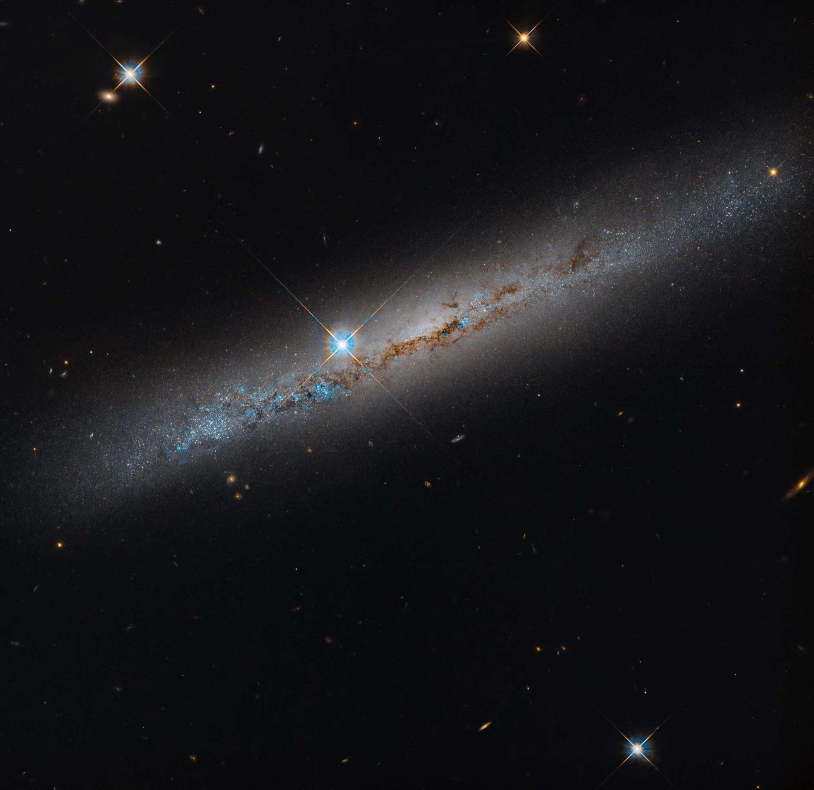 An edge-on spiral galaxy. Its rusty-brown dust lanes bisect the galaxy lengthwise. Bright-blue points dot the galaxy and seem to be more dense on its left side. A bright foreground star shines just to the left of the galaxy's center. The background is black and dotted with distant galaxies and a smattering of foreground stars, which are easily identified by their diffraction spikes.
