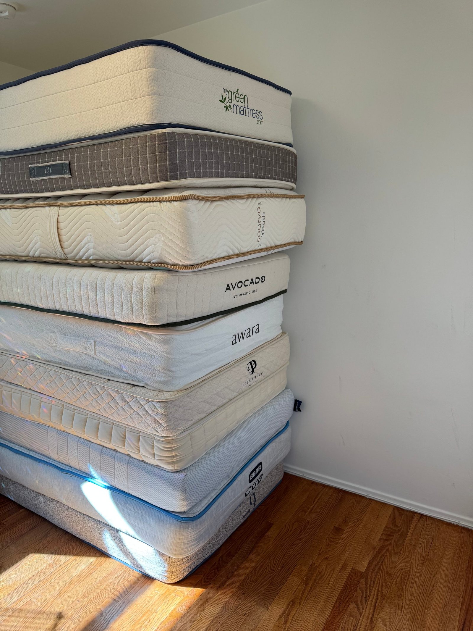 A stack of eight different brand-name mattresses against a white wall on a wooden floor.