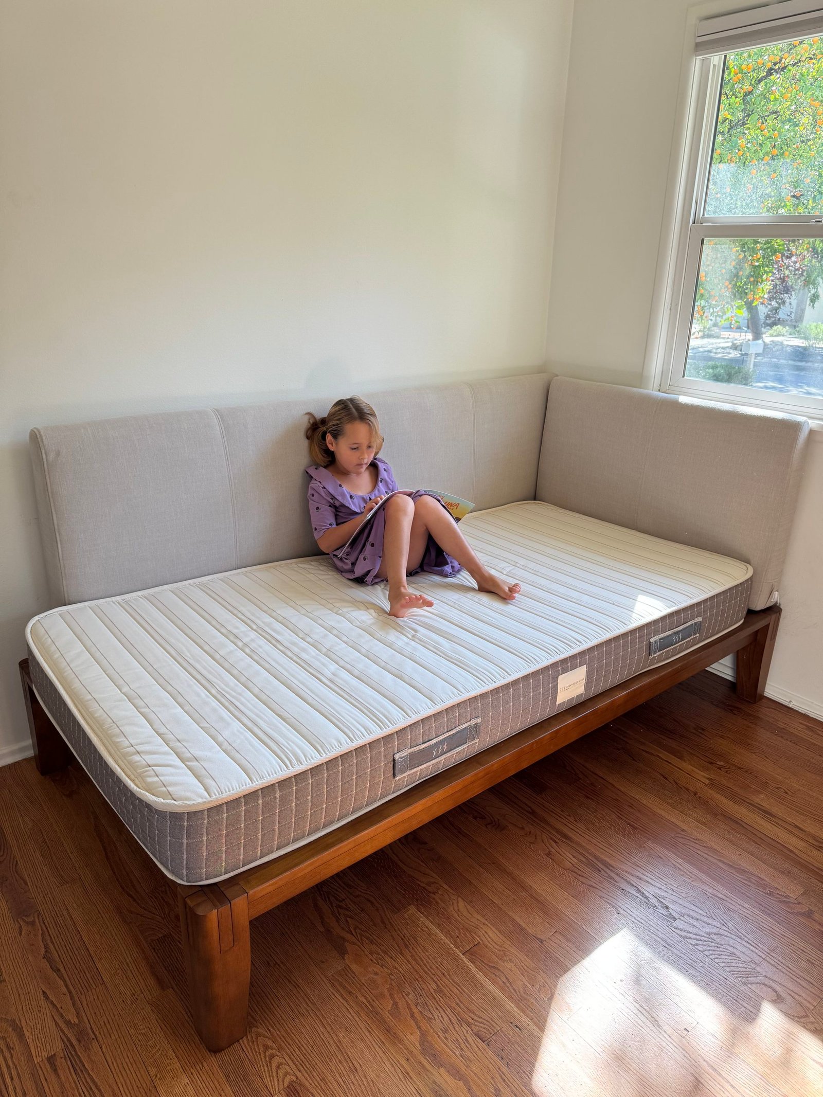 A young girl in a purple dress sits on a twin-sized mattress on a wooden frame, reading a book.