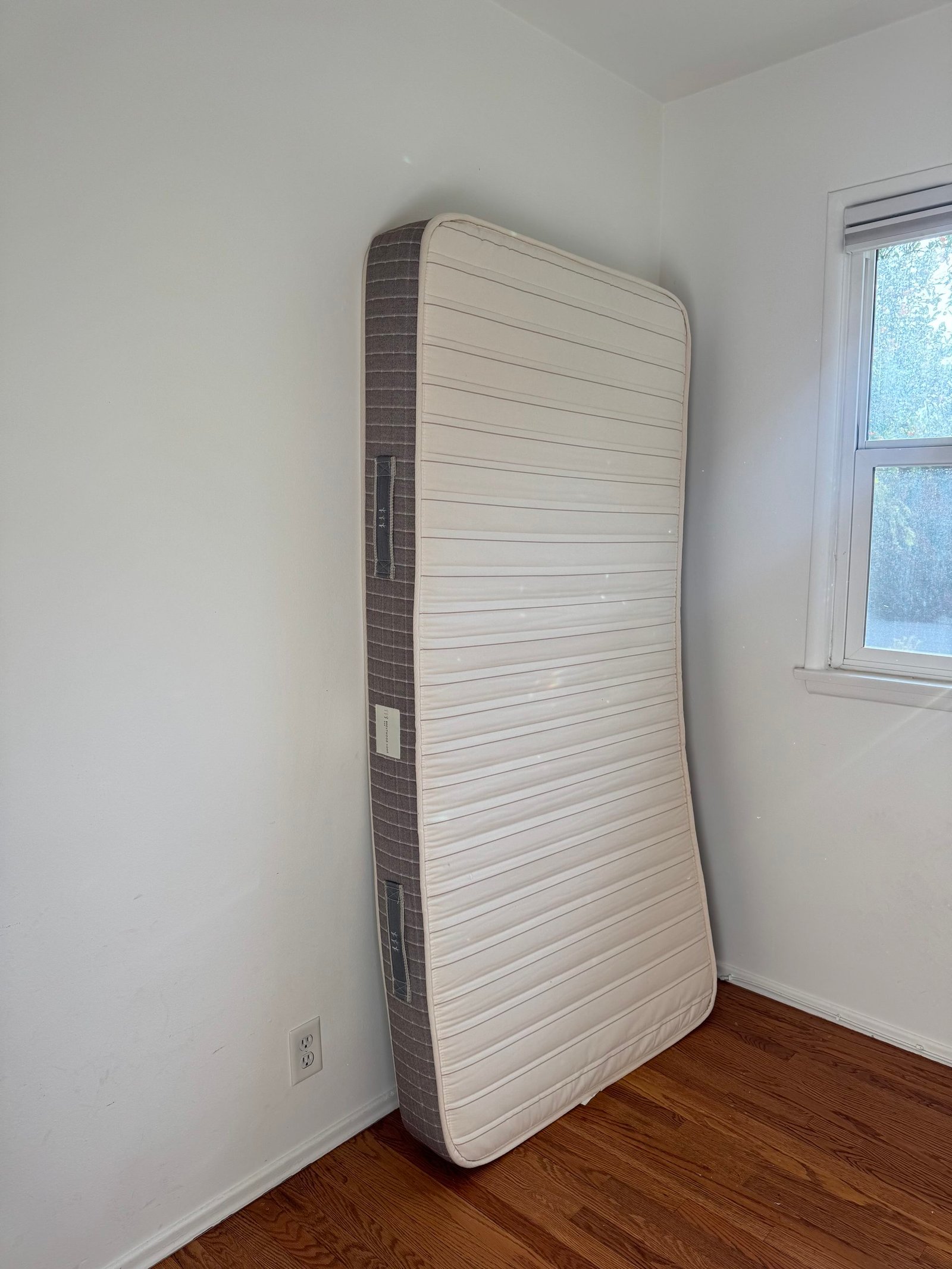 A mattress leaned against a white corner wall next to a window.