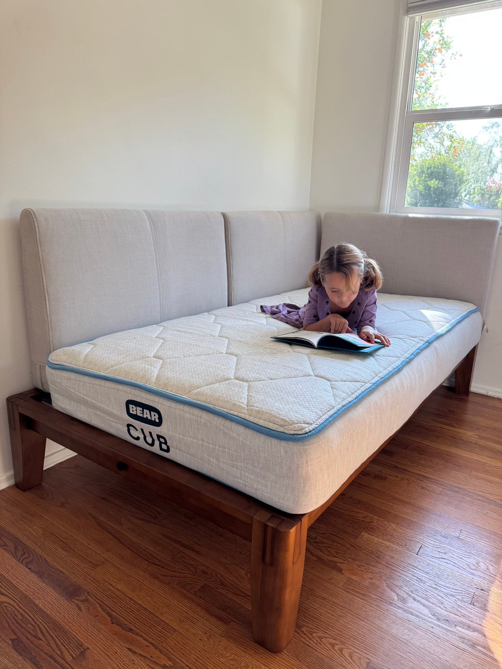 A young girl lying on a Bear Cub mattress on a wooden daybed, reading an open book.