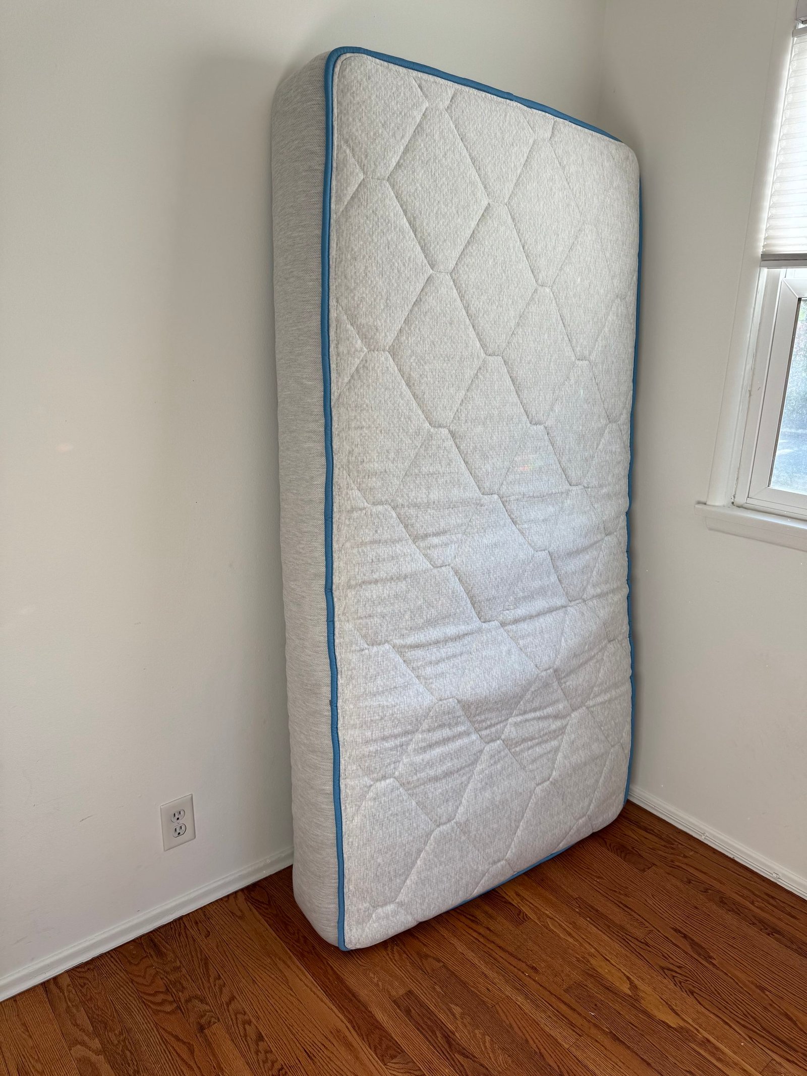A twin mattress with a quilted top and blue trim standing upright against a white wall.