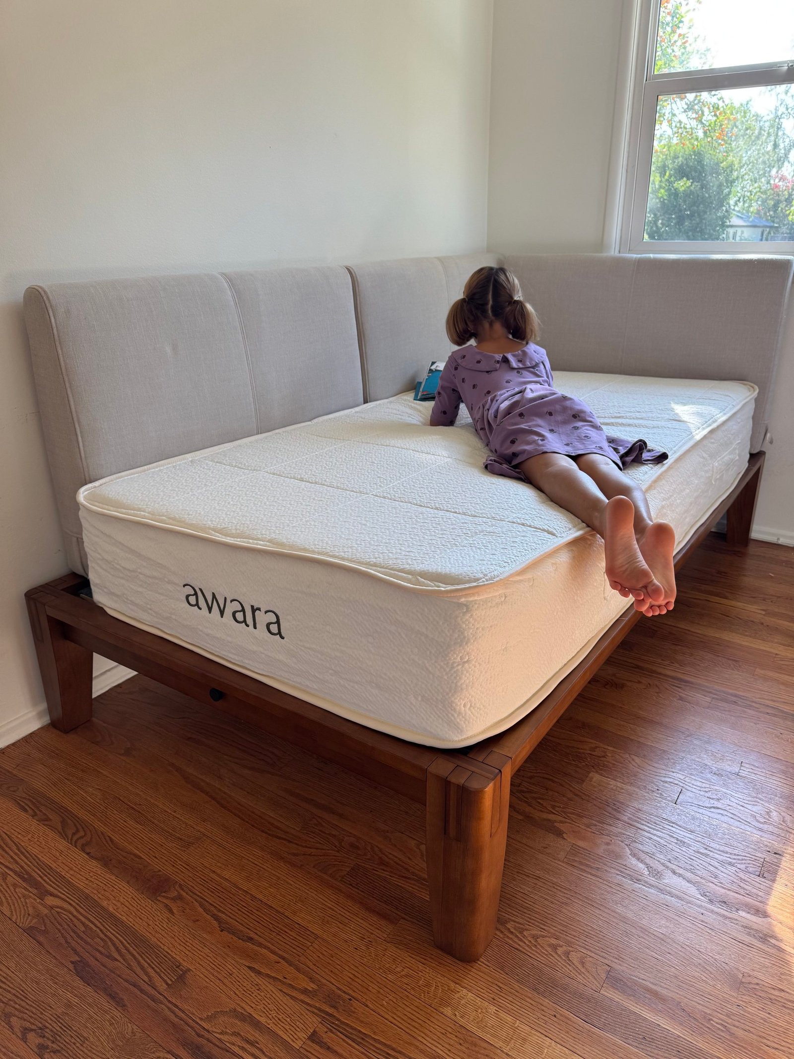 A child lies on their stomach reading a book on an Awara mattress placed on a wooden bed frame with an upholstered headboard.