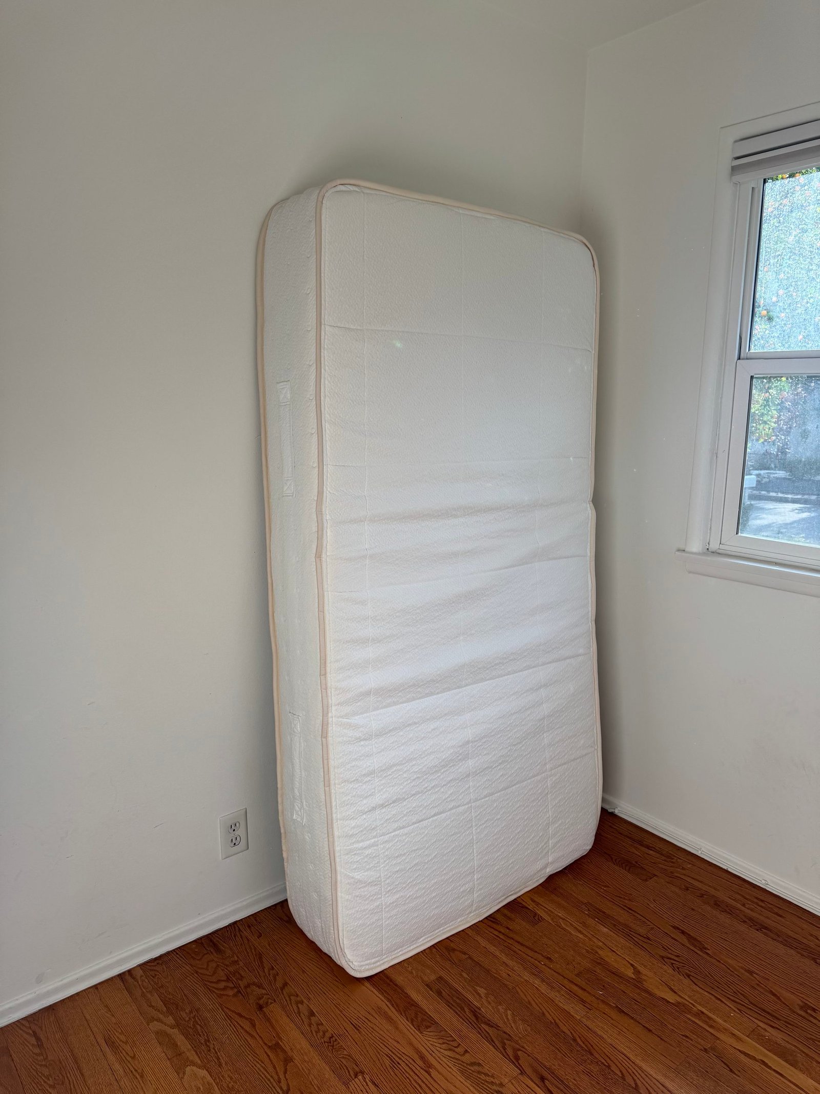 A white mattress standing upright in the corner of a room with hardwood floors.