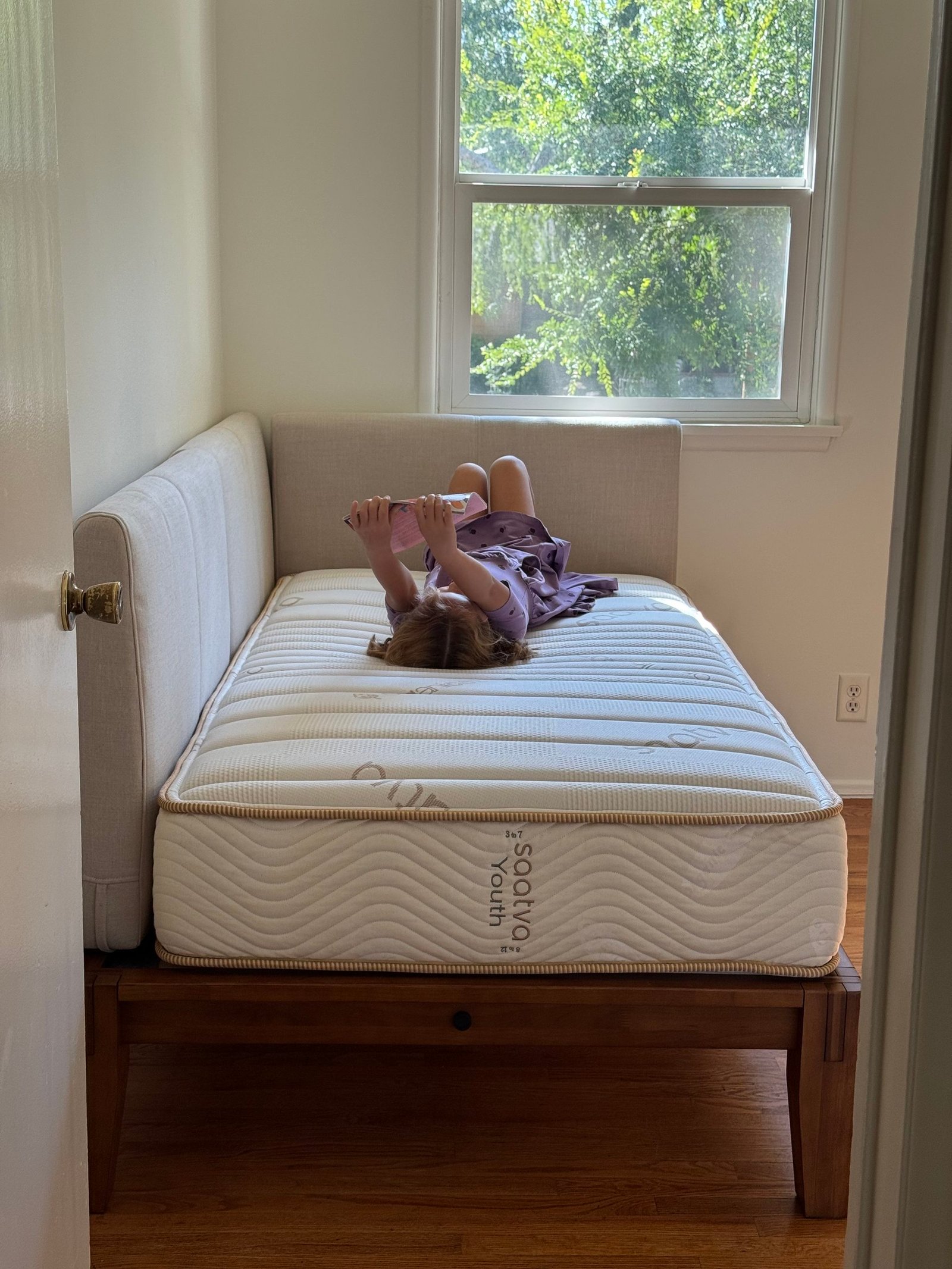 A child in a purple dress lies on their stomach on a Saatva youth mattress, reading a book.