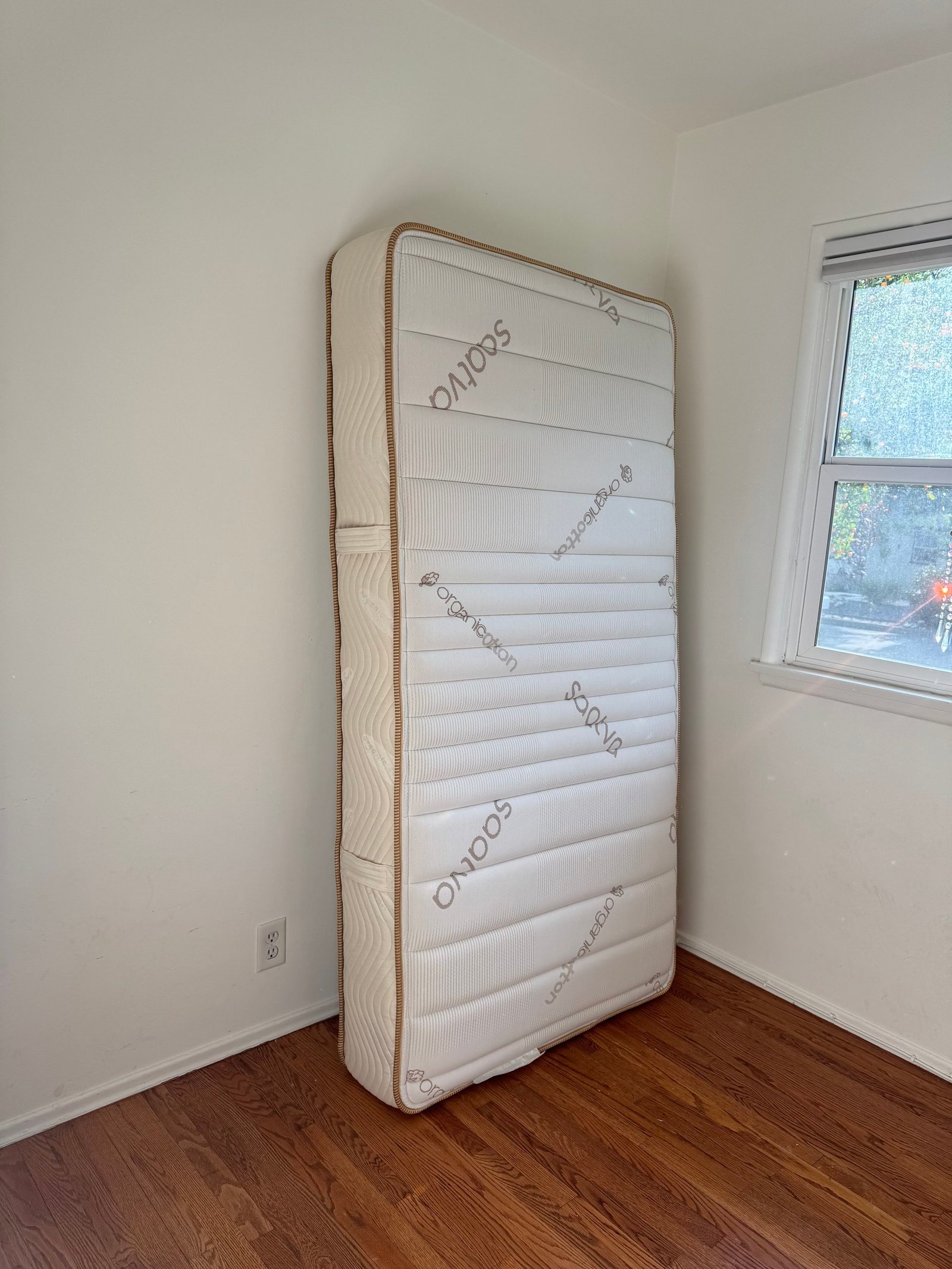 A Saatva organic cotton mattress standing upright against a white wall in the corner of a room with hardwood floors.