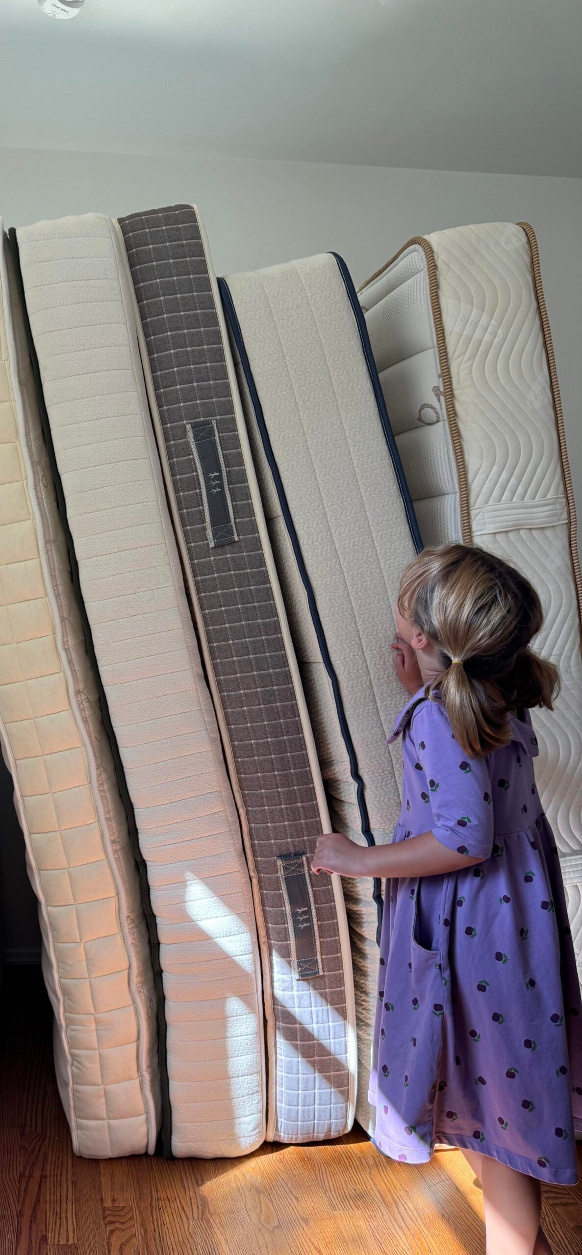 A child in a purple dress standing next to a row of several mattresses stood on their sides against a wall.