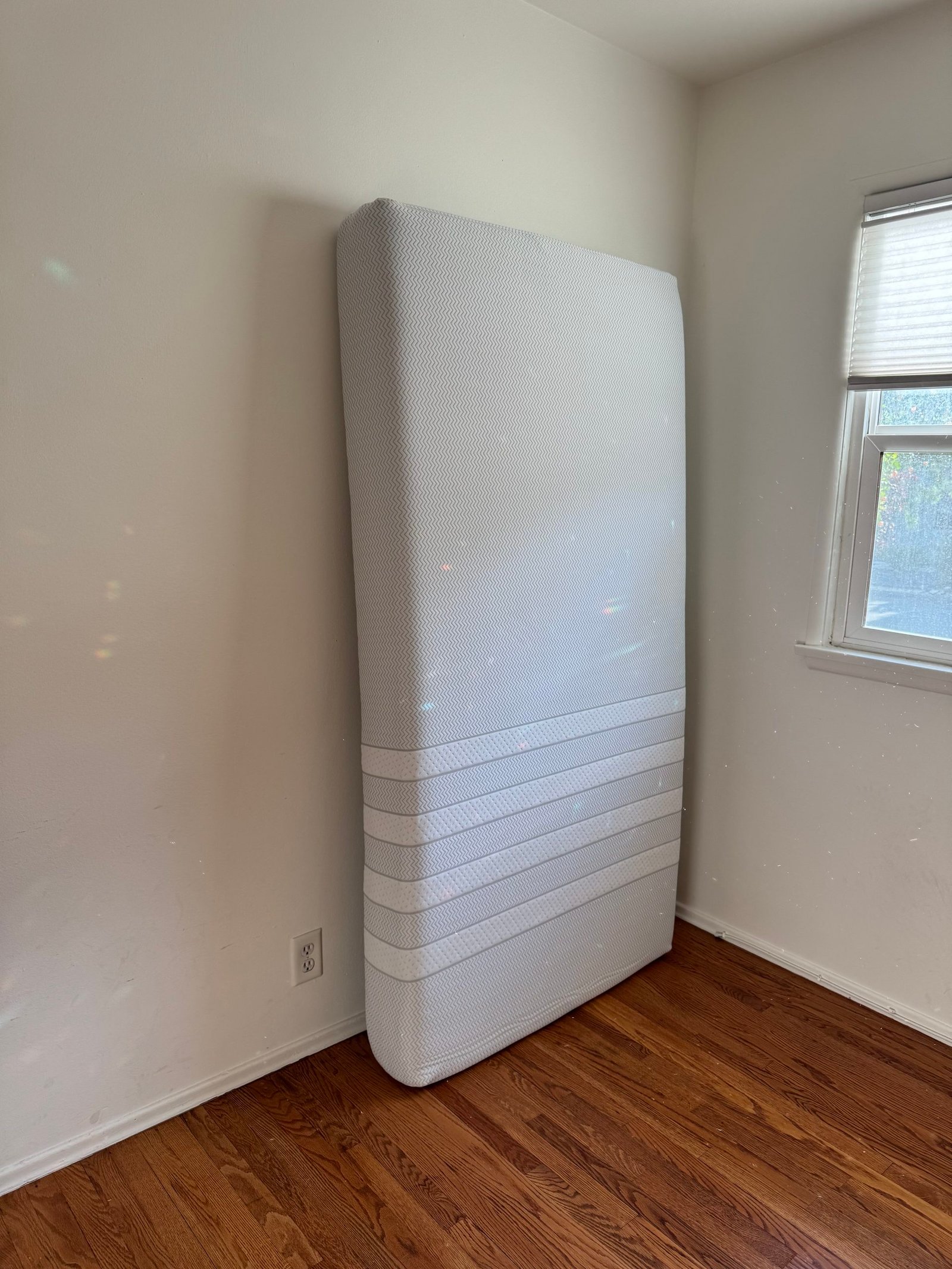 A white mattress standing upright in the corner of a room with hardwood floors.
