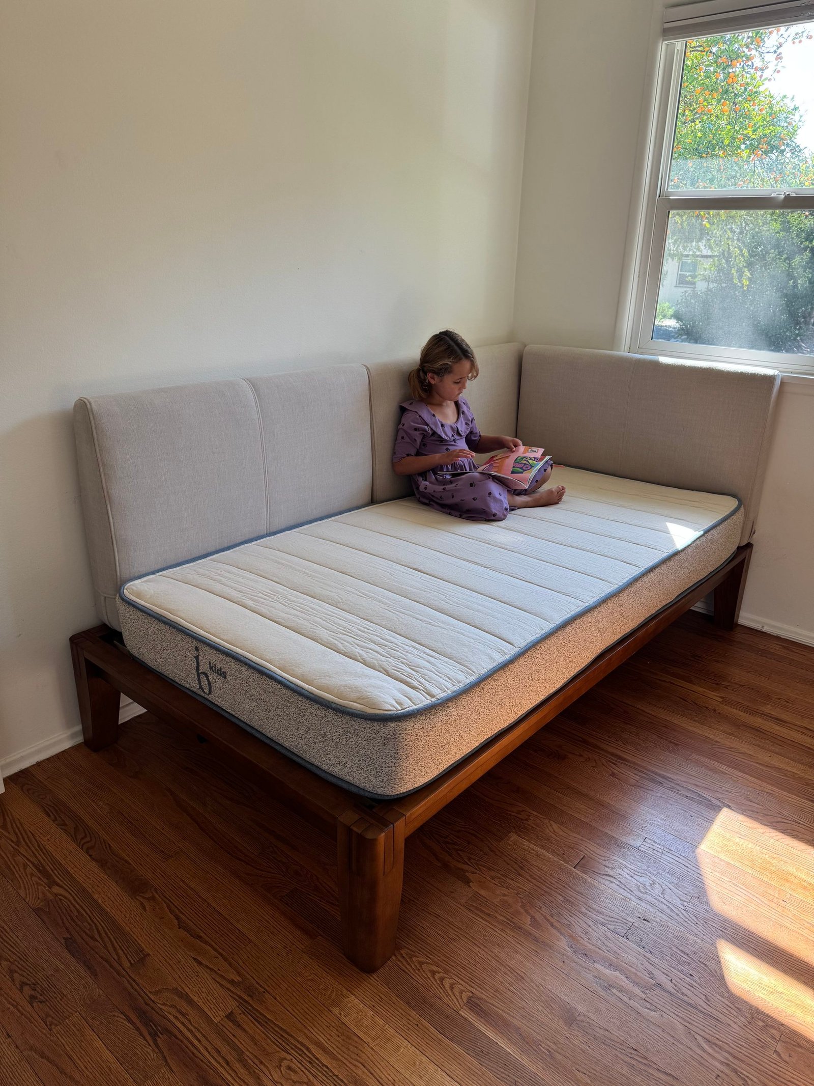 A child in a purple dress sits on a corner daybed reading a book.