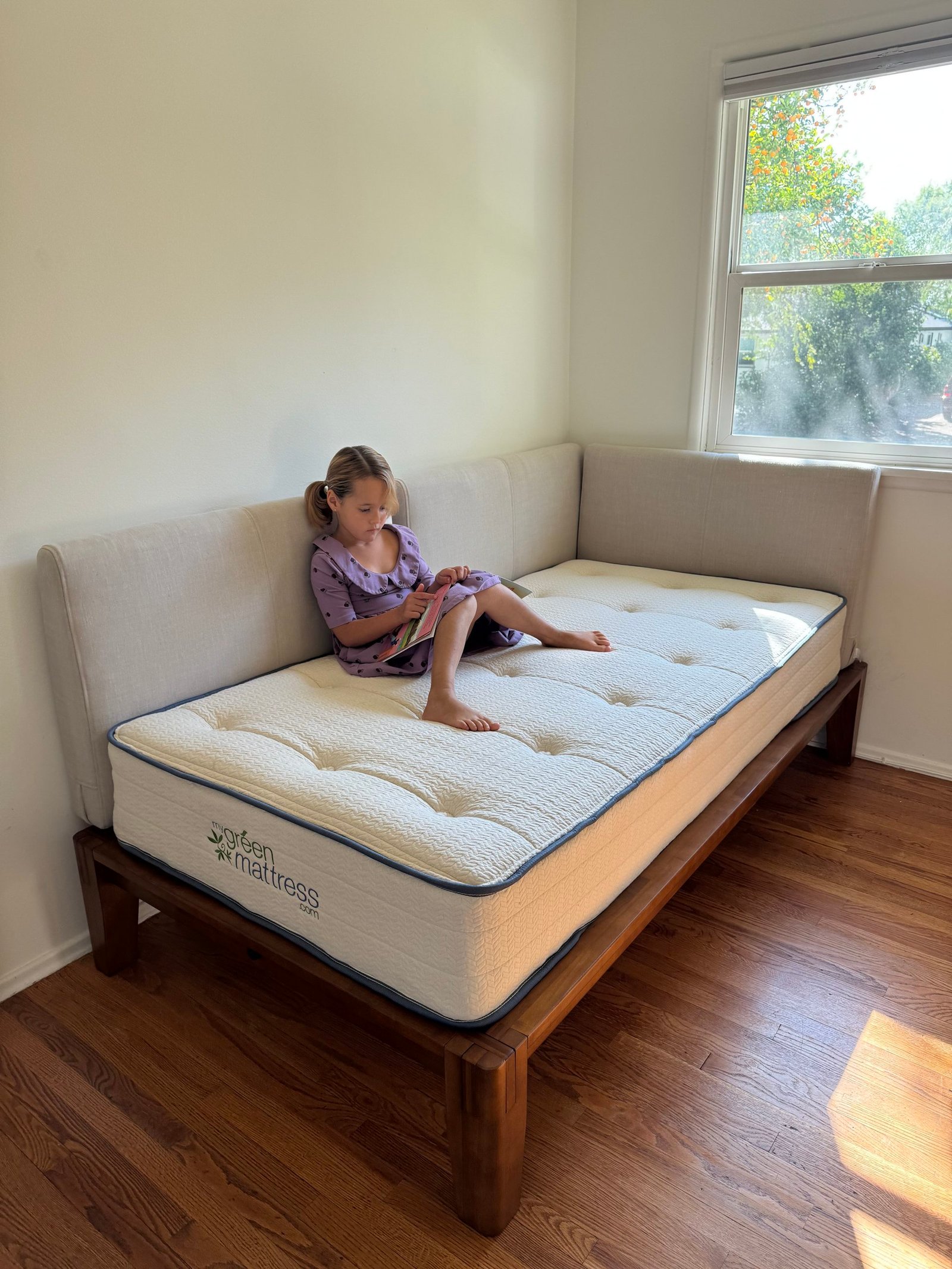 A young girl sitting on a mattress on a wooden frame, reading a book.