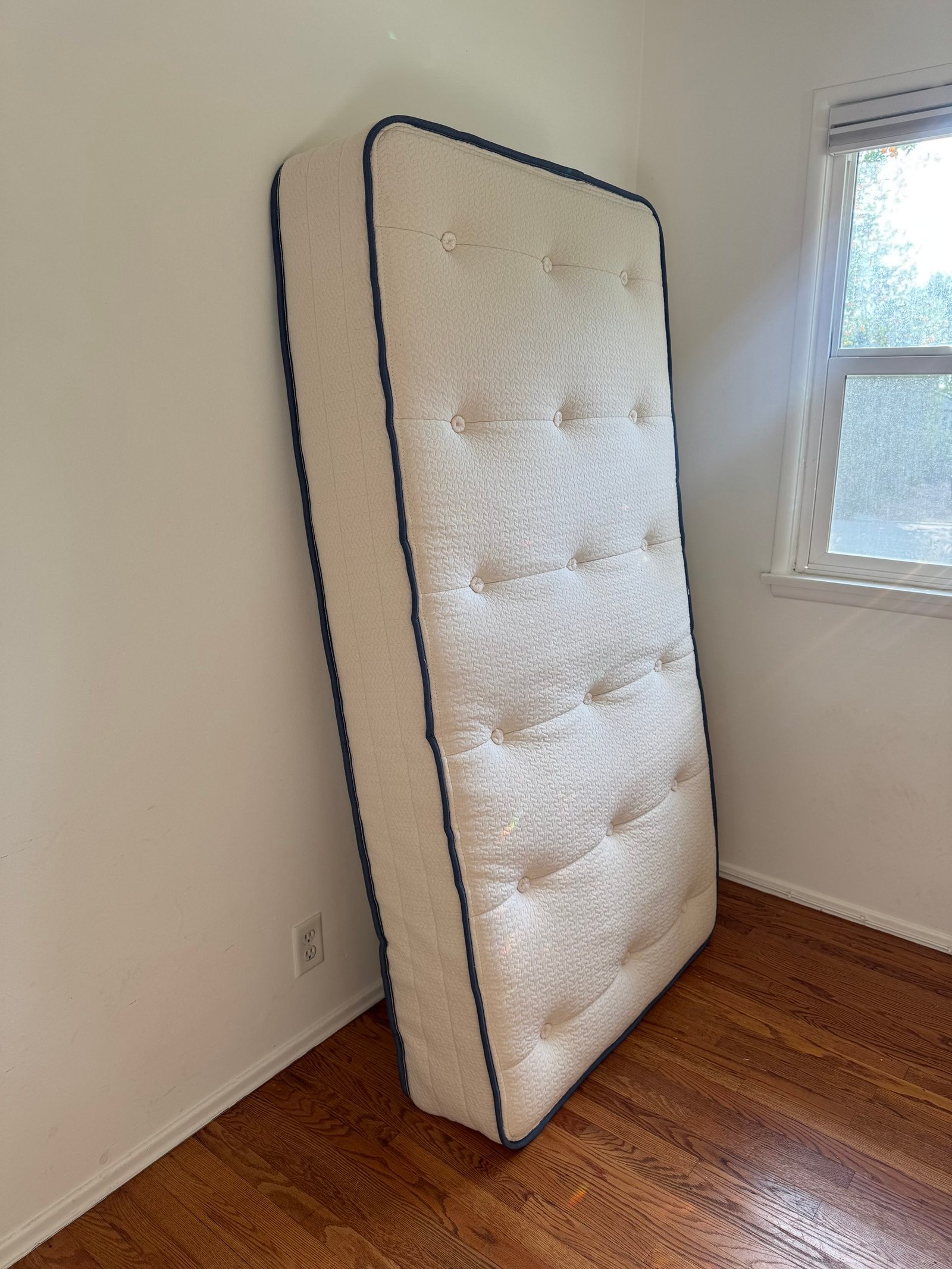 A twin-sized tufted mattress with blue piping, propped upright against a wall in a room.