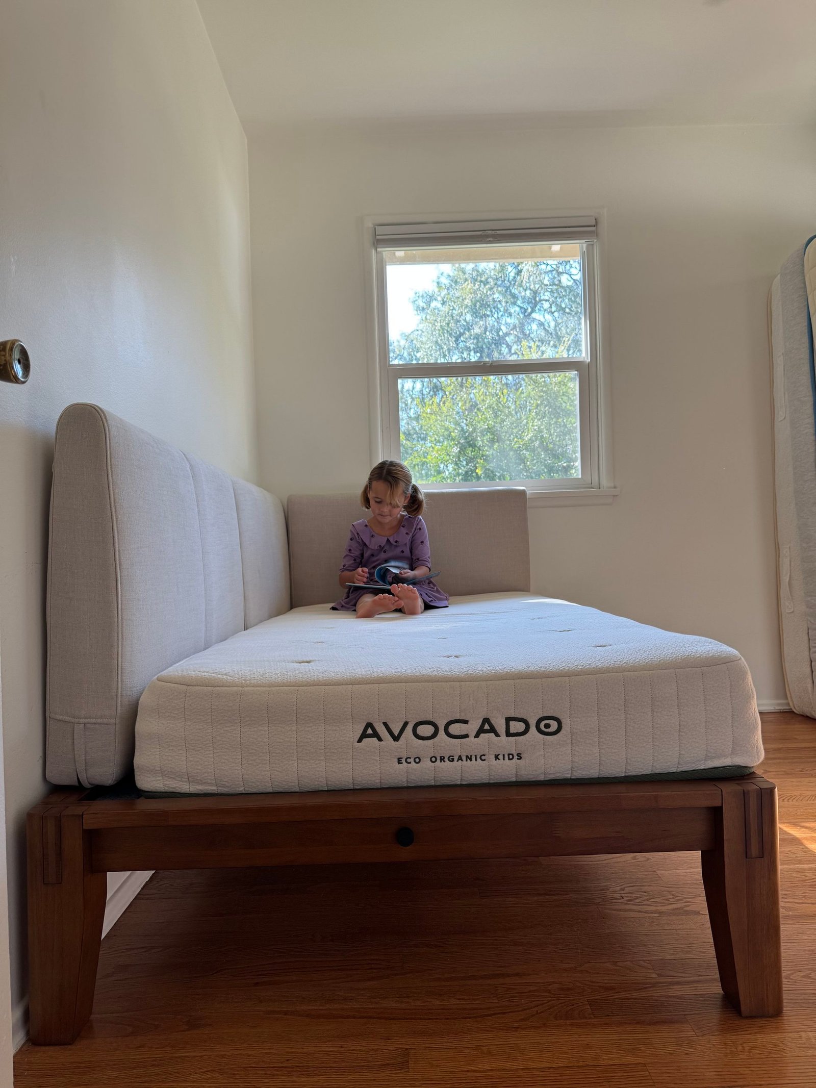 A child sitting on an Avocado Eco Organic Kids mattress on a wooden bed frame, reading a book.