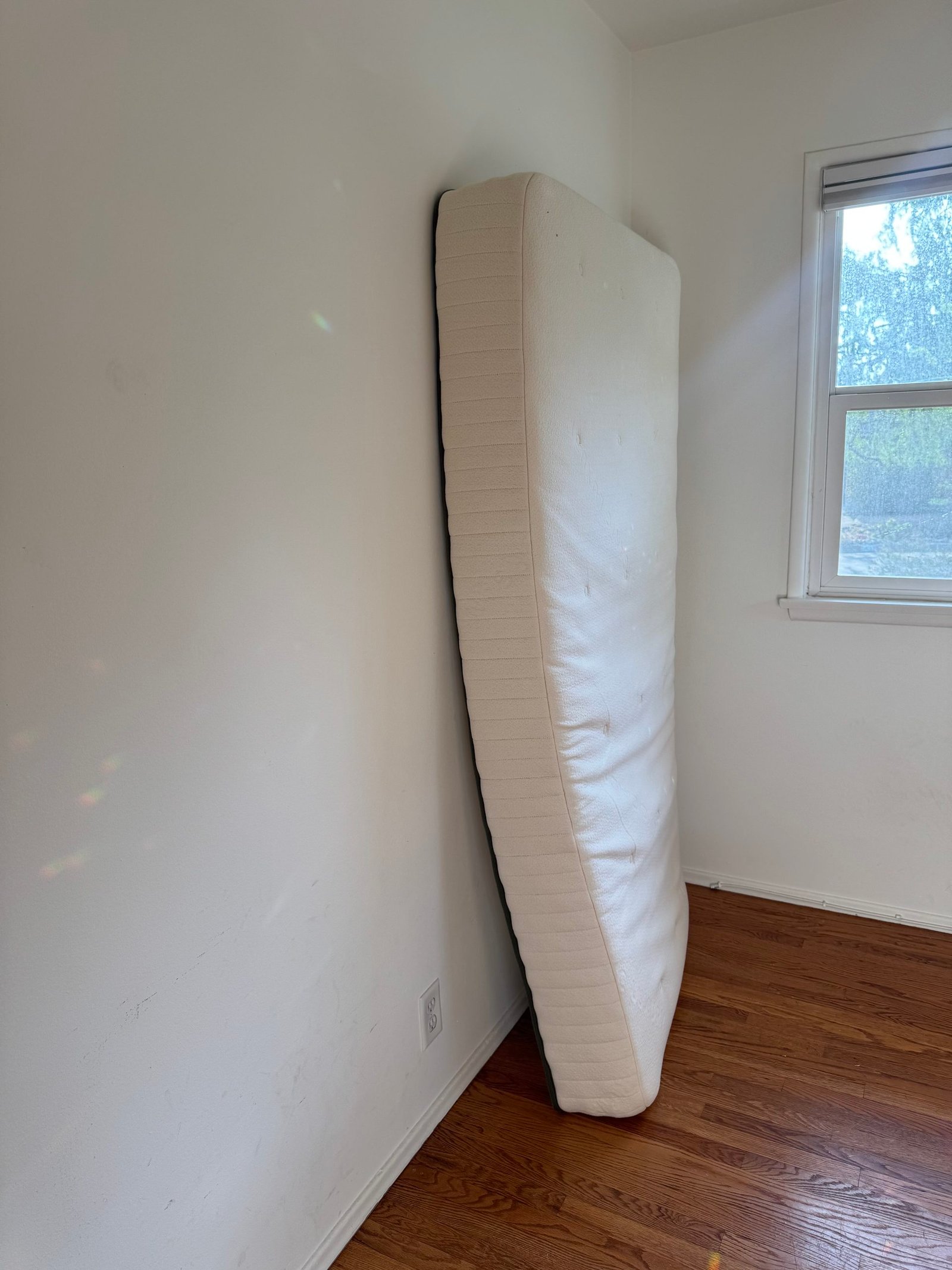 A mattress leaned upright against a white wall in a room with hardwood floors.