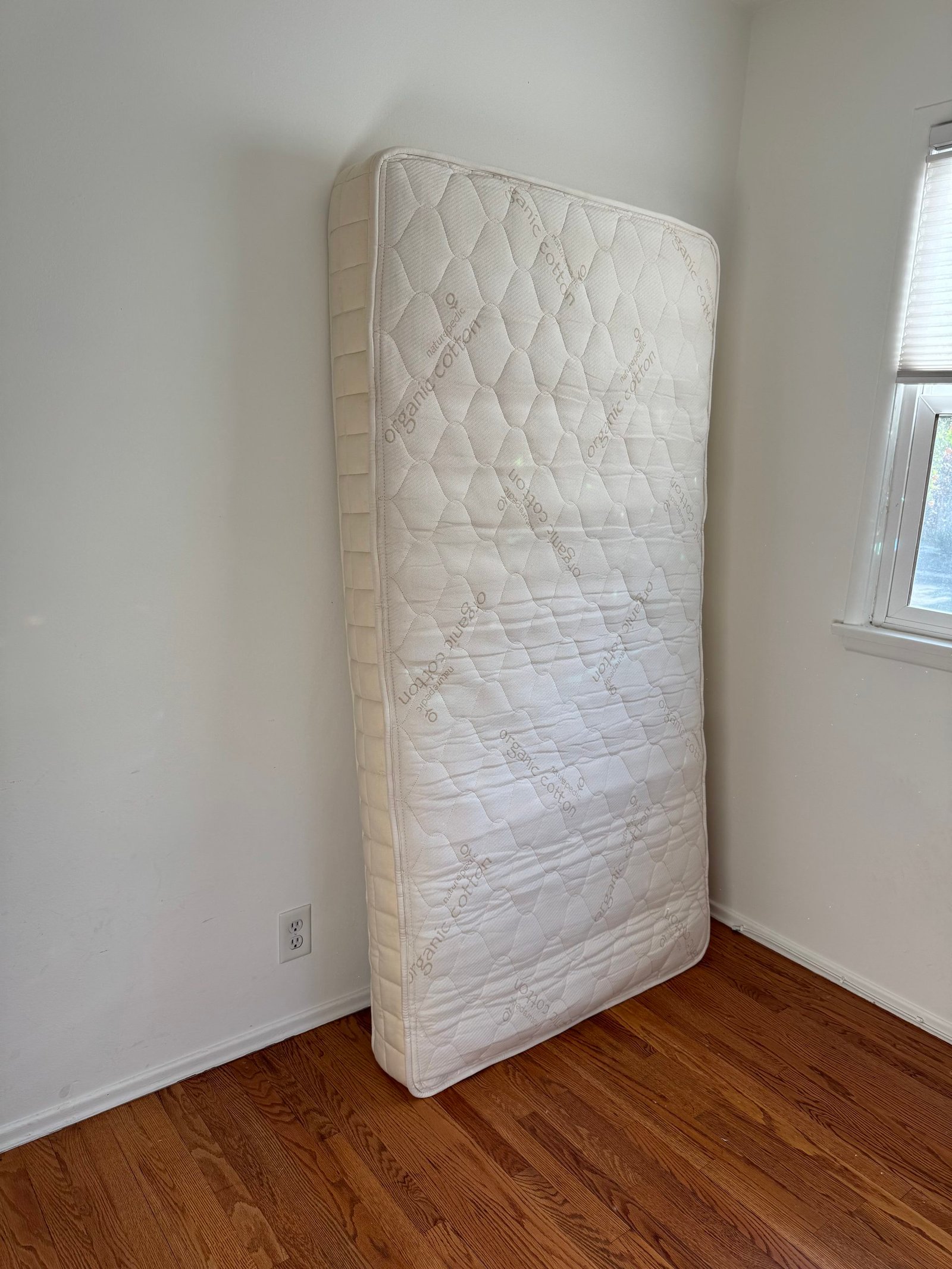 A twin mattress standing upright against a white wall on a wooden floor.