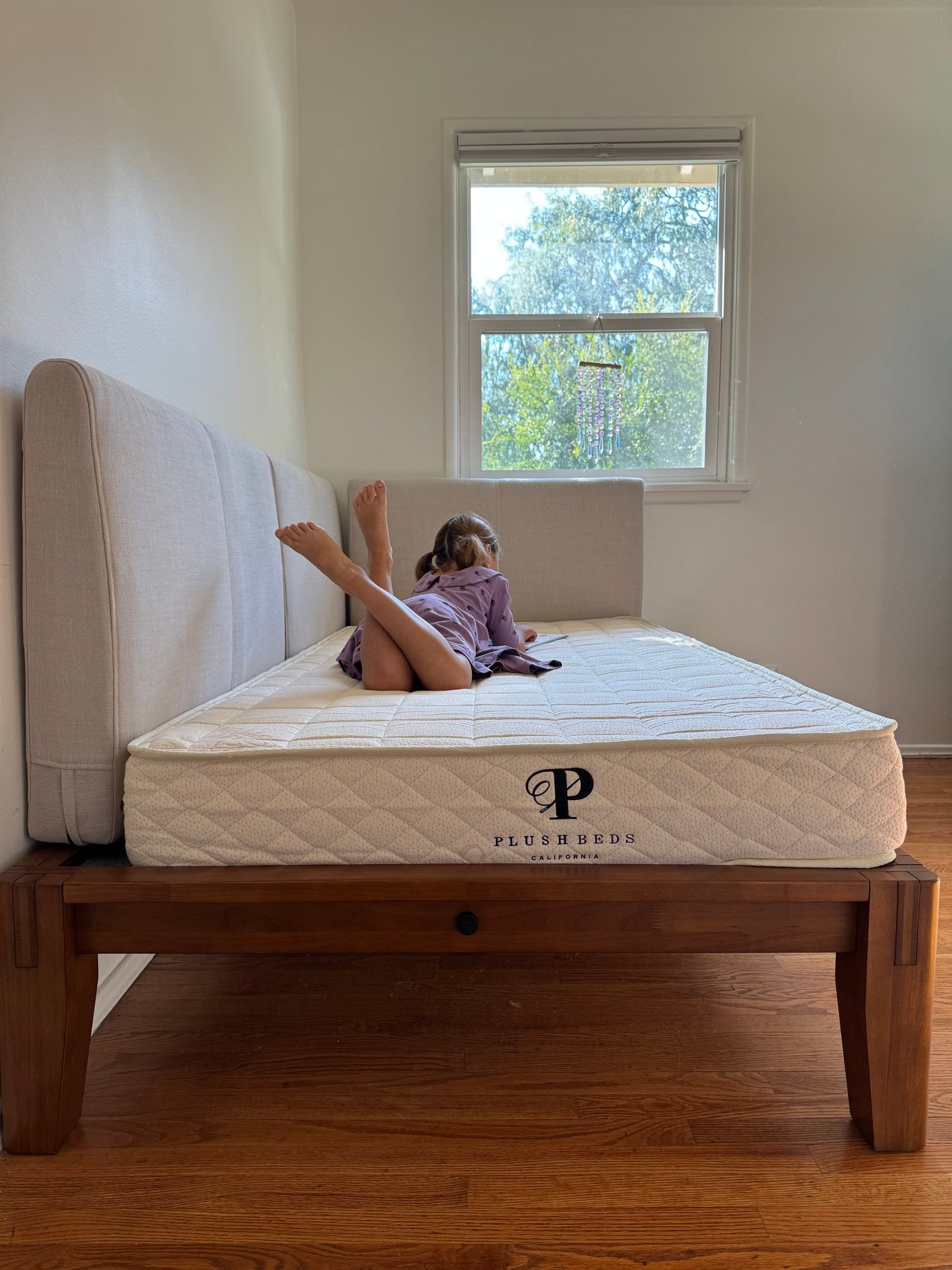 A child lies on their stomach reading on a PlushBeds mattress atop a wooden bed frame.