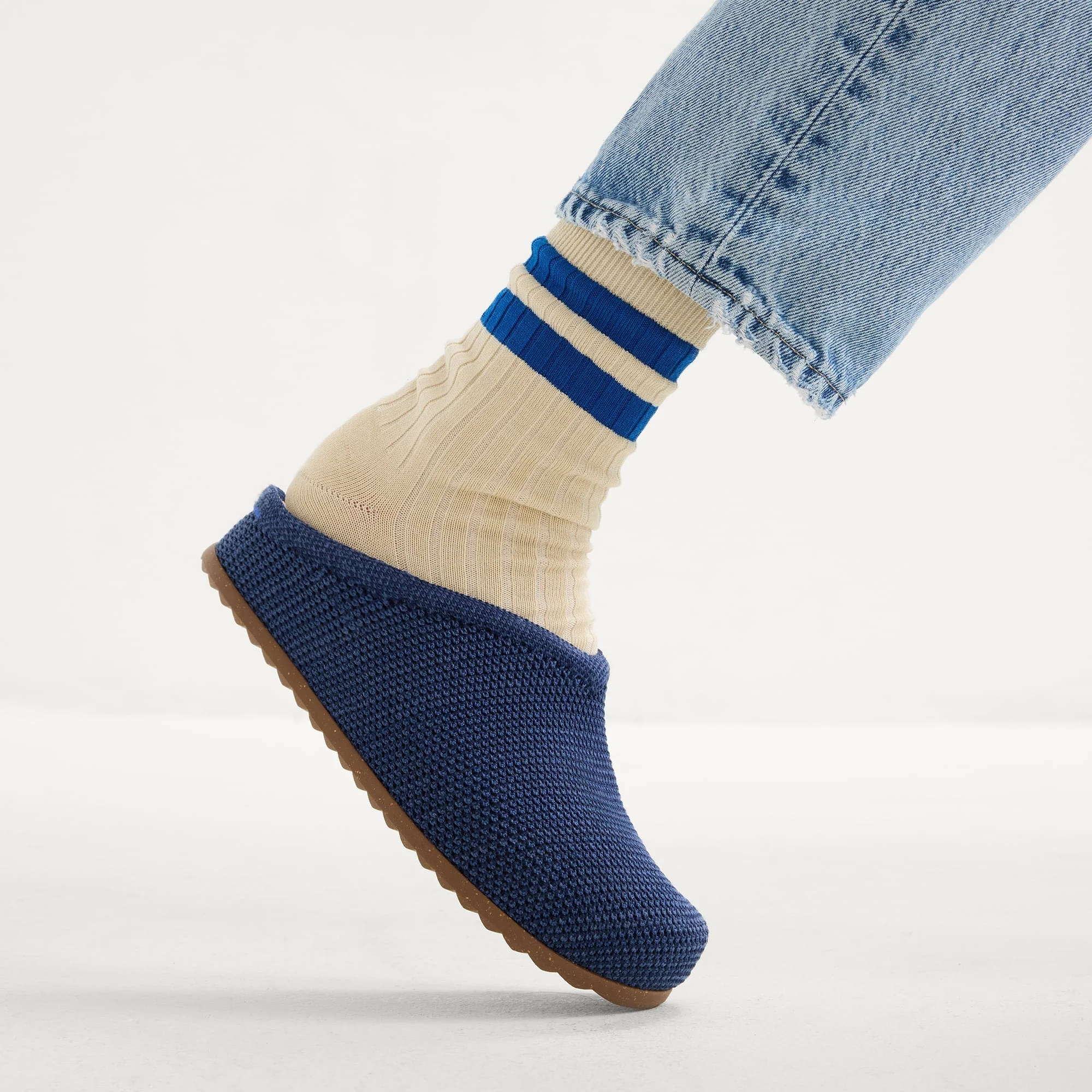 A person wearing cream ribbed socks with blue stripes, blue knit slippers, and light blue jeans, standing on a white surface.