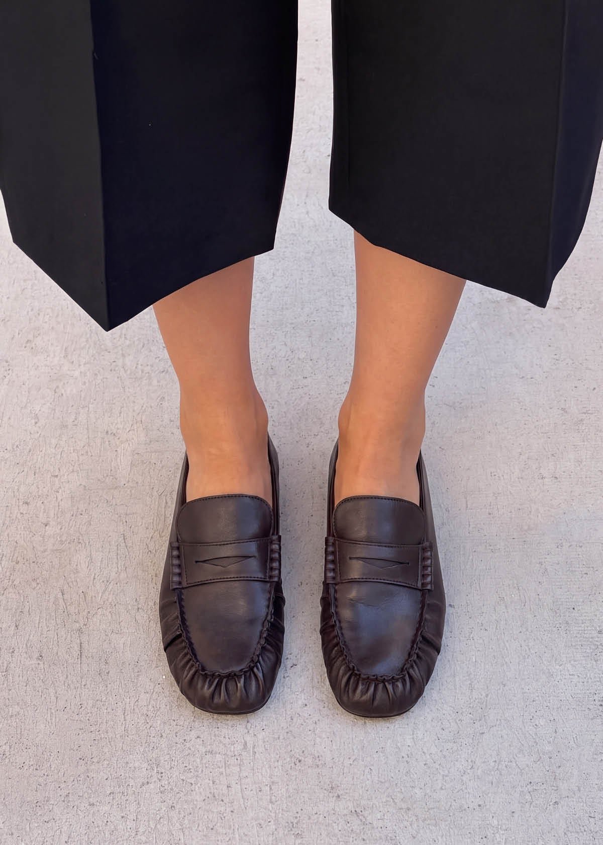A person wearing black wide-leg trousers and dark brown leather penny loafers.